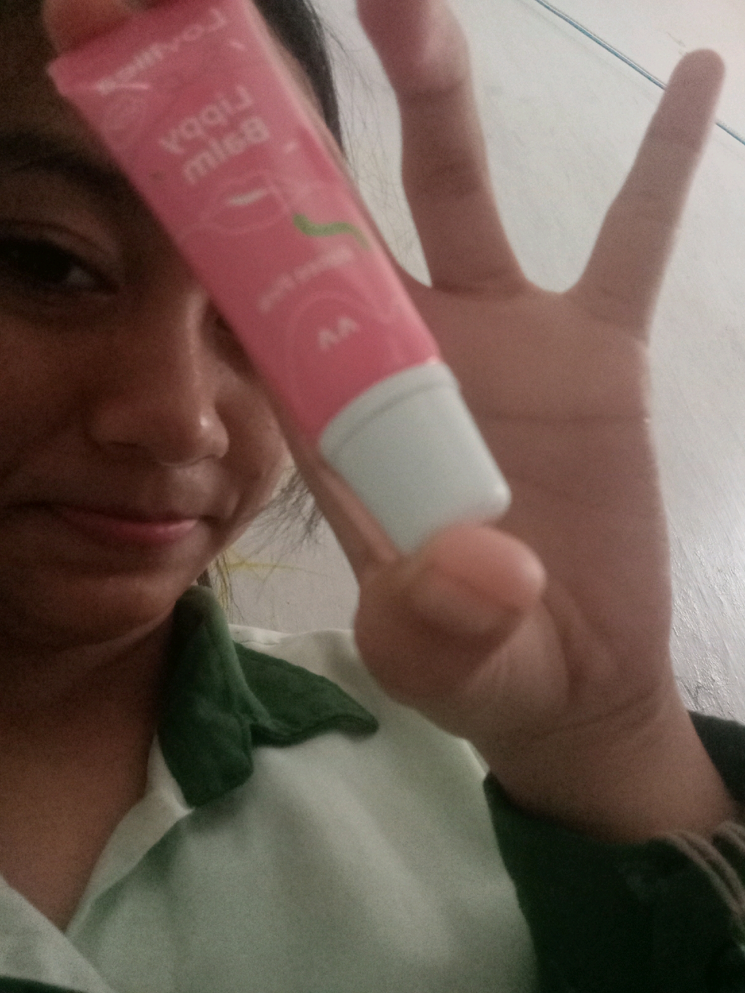 A person, likely a young female, is holding a pink lip balm tube in front of their face. The person is wearing a white collared shirt with dark green accents on the collar and sleeves. The background appears to be a lightly colored, textured wall. The image was taken indoors. The location of Badas, Indonesia, is not discernible from the provided image.