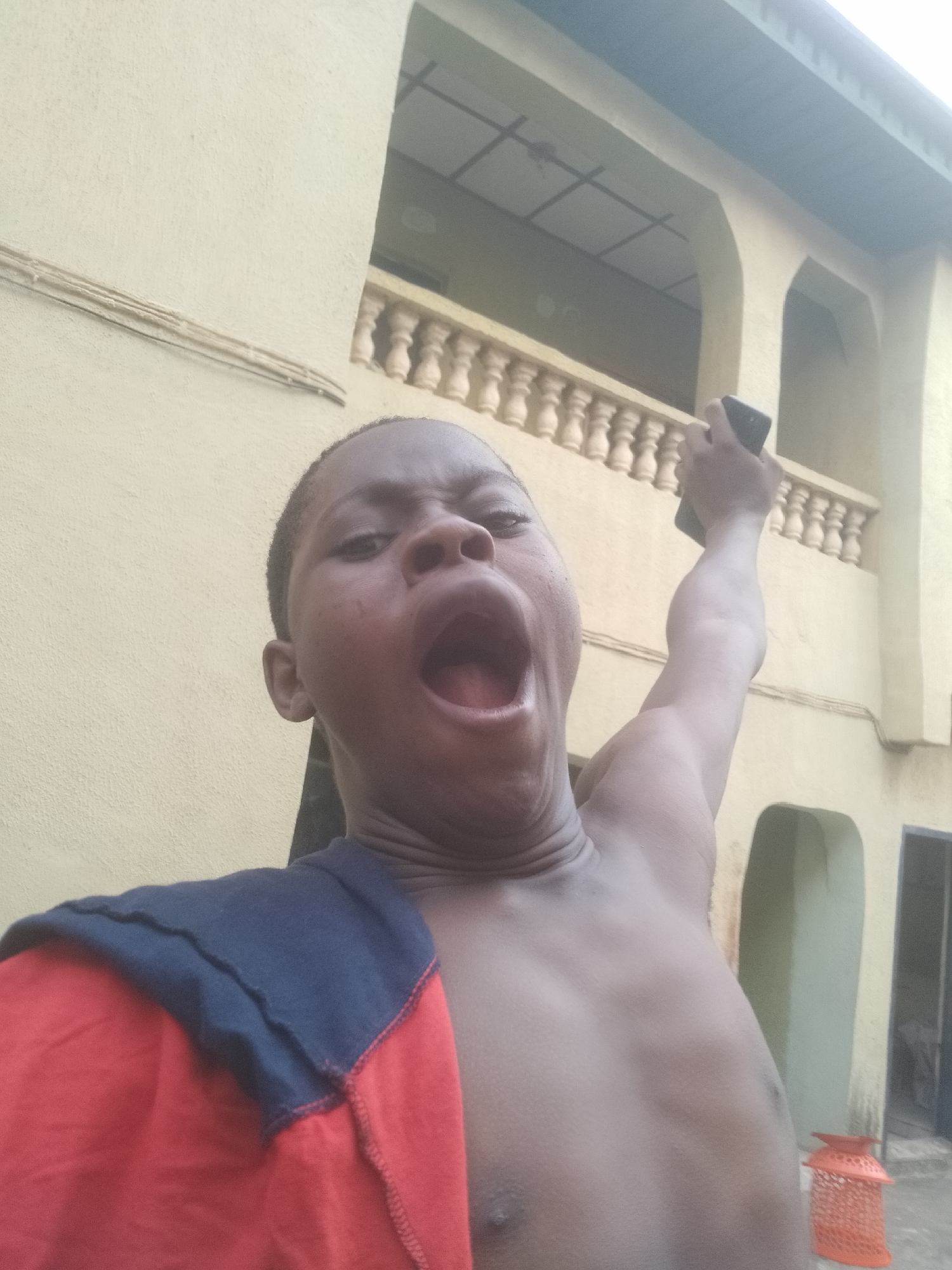 A shirtless person with dark skin is yawning, holding a black rectangular object in their raised right hand, possibly a phone, against the backdrop of a light-colored building with arches and a balustrade. The person is wearing a red and blue garment draped over their left shoulder. The location is a city of Unknown, Unknown.