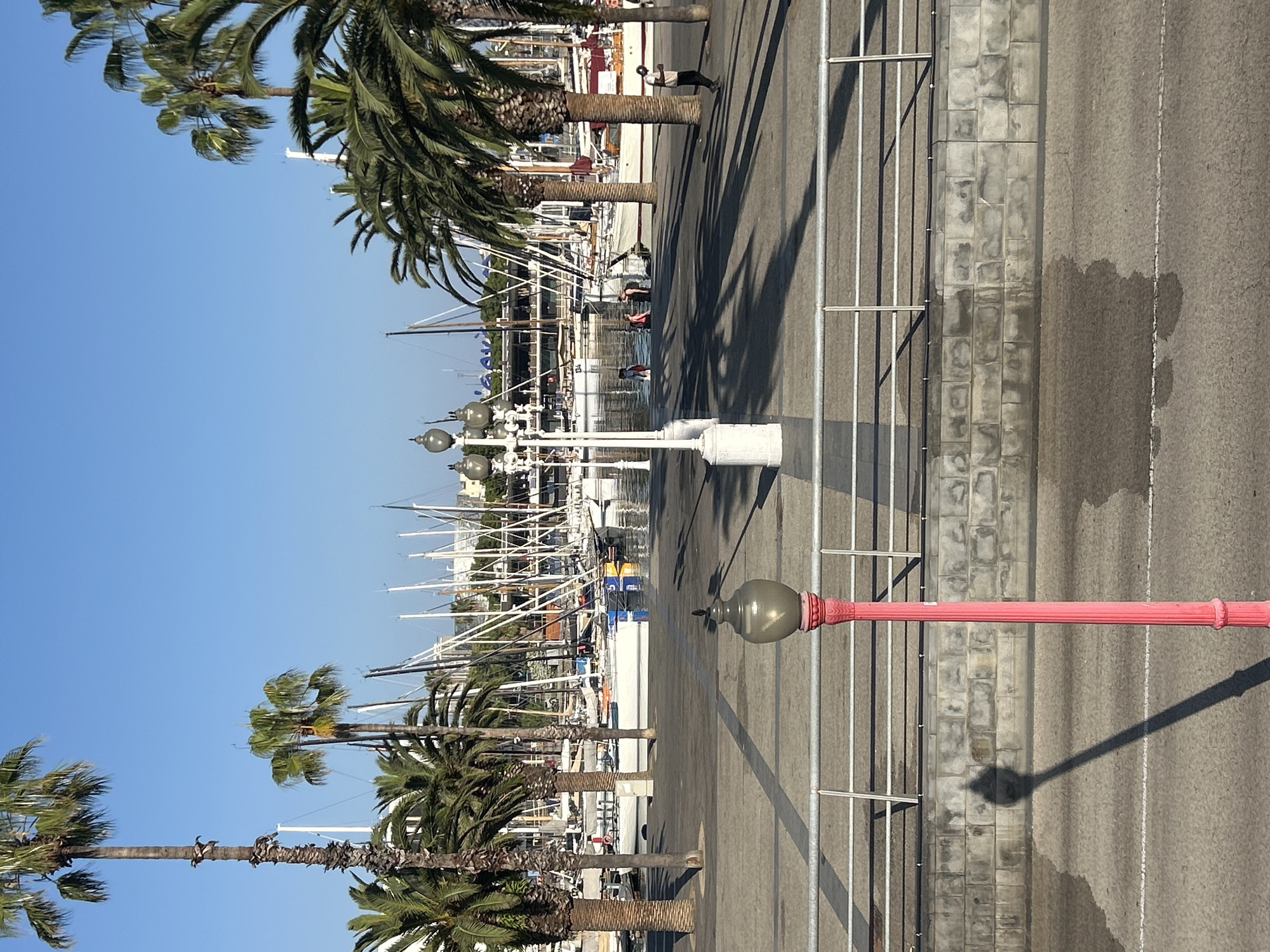 This image depicts a sunny day at a harbor in Barcelona, Spain. The scene is dominated by a clear blue sky and the masts of numerous sailboats lining the waterfront. Palm trees with lush green fronds cast long shadows across the paved area. In the foreground, a bright pink lamppost with a glass globe stands tall, its shadow stretching across the pavement.
Along the waterfront, several people are visible. One individual is leaning against a railing, looking out at the boats, while others can be seen walking or standing near the water's edge. A person is captured mid-stride on the paved walkway. In the distance, more figures can be seen near the boats.
The boats themselves are a prominent feature, with their white hulls and tall masts creating a striking visual pattern against the backdrop of the city skyline. The water of the harbor is calm, reflecting the clear sky. The pavement appears well-maintained, with white lines marking lanes and a textured stone border along the edge of the harbor. The overall atmosphere is one of leisure and a bright, warm day. The presence of palm trees and the style of the boats are strong indicators of a Mediterranean location.
