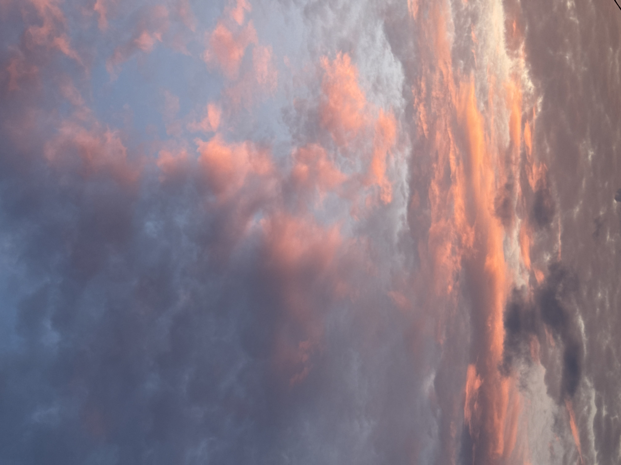The image shows a sky filled with dramatic clouds, painted in hues of pink, orange, and deep blue. The clouds are wispy and textured, with some areas appearing lighter and more ethereal, while others are darker and more substantial. The lighting suggests a sunset or sunrise, casting a warm glow on the upper parts of the clouds. The lower portion of the image is dominated by darker, gray clouds, creating a contrast with the vibrant colors above. The scene is devoid of any identifiable landmarks or structures, suggesting an open sky view, possibly from an outdoor location. The specific location in Бараўлянскі сельскі Савет, Belarus, is not visually discernible from the image itself.