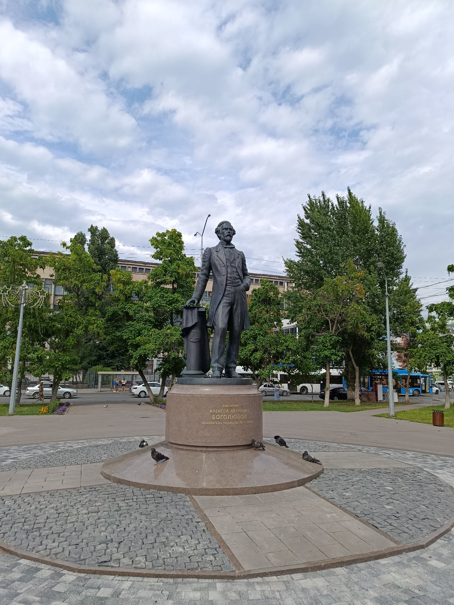 The image shows a full shot of a bronze statue of a man in a suit, standing on a stone pedestal. The pedestal has inscriptions in Russian. The statue is located in a public space with paved walkways and cobblestone areas. Pigeons are present on the ground around the base of the statue. In the background, there are trees, buildings, a street with cars and a blue bus, and a cloudy sky. The inscription on the pedestal reads "ХУДОЖНИК АЛЕКСЕЙ ПЕТРОВИЧ БОГОЛЮБОВ ОСНОВАТЕЛЬ САРАТОВСКОЙ АКАДЕМИИ ХУДОЖЕСТВ" which translates to "ARTIST ALEXEY PETROVICH BOGOLYUBOV FOUNDER OF THE SARATOV ACADEMY OF ARTS". This indicates the statue is located in Saratov, Russia.