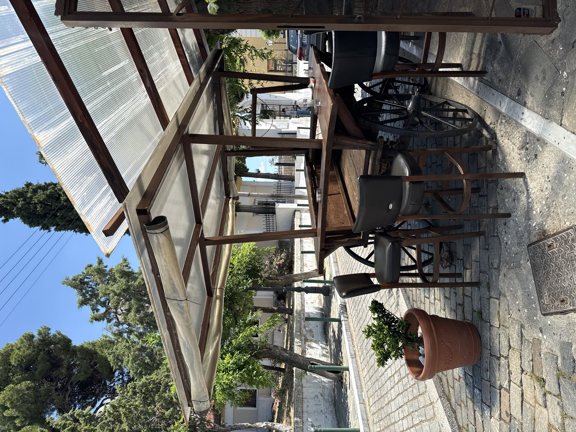 The image captures an outdoor seating area in Kavala, Greece, featuring a unique bar constructed from a wooden wagon. The wagon bar is equipped with a wooden countertop and several dark-upholstered bar stools. A transparent corrugated plastic roof, supported by a wooden and metal frame, covers the seating area, providing shade.
In the foreground, a potted plant with green foliage sits on a cobblestone pavement. The street is paved with gray cobblestones and bordered by a sidewalk. Behind the wagon bar, a row of trees with lush green foliage is visible, and further in the background, buildings with white and yellow facades, characteristic of Mediterranean architecture, are present. A dark-colored car with a license plate is parked on the street. The sky is clear and blue, suggesting a sunny day.
The scene appears to be a commercial establishment, possibly a cafe or bar, designed to offer an outdoor dining or drinking experience with a rustic, charming aesthetic. The overall ambiance is relaxed and inviting, suitable for a warm, sunny day.