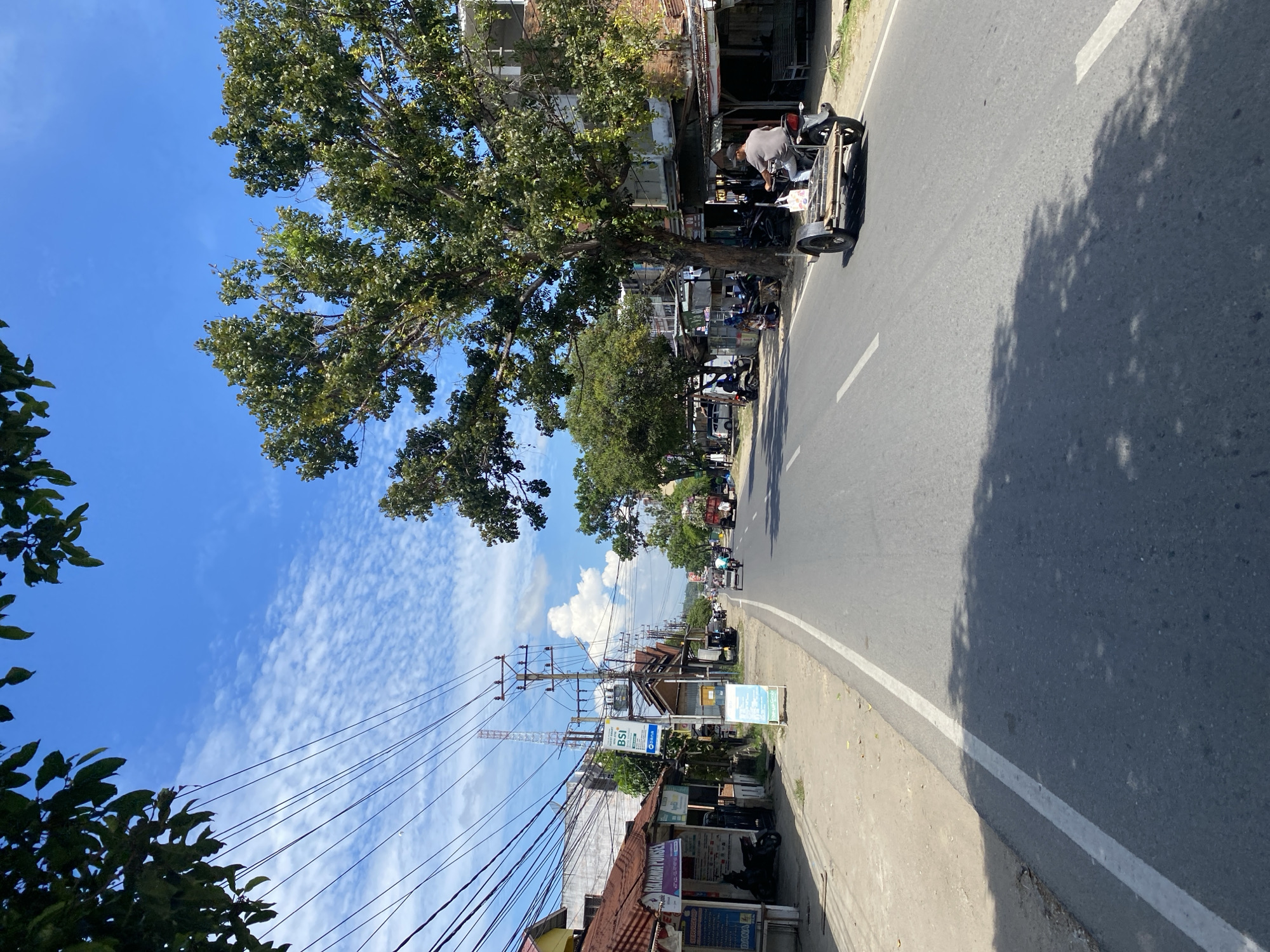 The image depicts a street scene in Lhokseumawe, Indonesia. The viewpoint is from the roadside, looking down the street. A large tree with green foliage dominates the left side of the frame, casting shadows on the asphalt road. The road has a white line marking the lane.
In the foreground, on the right side of the road, a person is working on a three-wheeled vehicle, possibly a motorcycle with a sidecar. Several other motorcycles are parked along the roadside in front of shops. The buildings lining the street appear to be a mix of commercial establishments with simple facades and corrugated metal roofs. Utility poles with a dense network of wires are prominent along the sidewalk.
The sky is a vibrant blue, dotted with scattered white clouds, suggesting a clear day. The sunlight is bright, indicated by the strong shadows. The presence of utility poles and the architecture of the buildings suggest a built-up urban or semi-urban environment.
Visible text includes "BSI" on a blue and white sign, likely referring to Bank Syariah Indonesia. Other signs are present but largely illegible due to resolution or distance. The overall atmosphere appears to be one of daily activity on a sunny day in a Southeast Asian locale.
