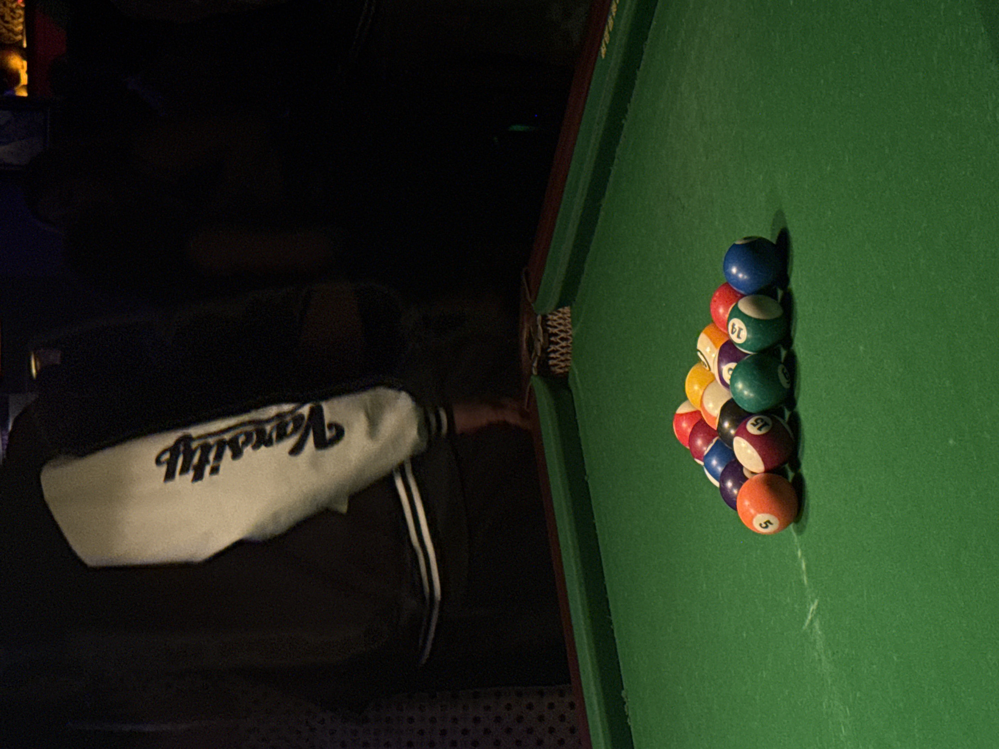 A person wearing a black jacket with a white "Varsity" print is standing next to a pool table. The pool table is covered in green felt. A rack of billiard balls is situated at one end of the table, ready to be broken. The balls are arranged in a triangular pattern. The setting appears to be indoors, possibly a bar or recreational area, as it's dimly lit. The image offers no visual evidence to confirm a location in Liangjiang, China.