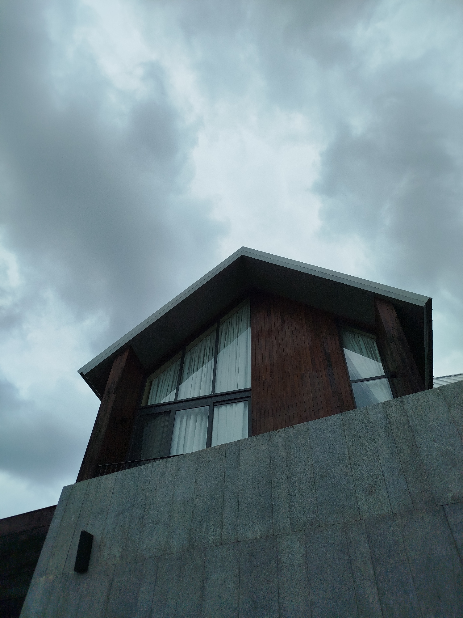 A low-angle, upward-facing shot of a modern building in Da Lat, Vietnam, under a cloudy sky. The building features a concrete base topped with a wooden structure with a triangular window. White curtains are visible behind the glass of the window. The sky is overcast with gray and white clouds.