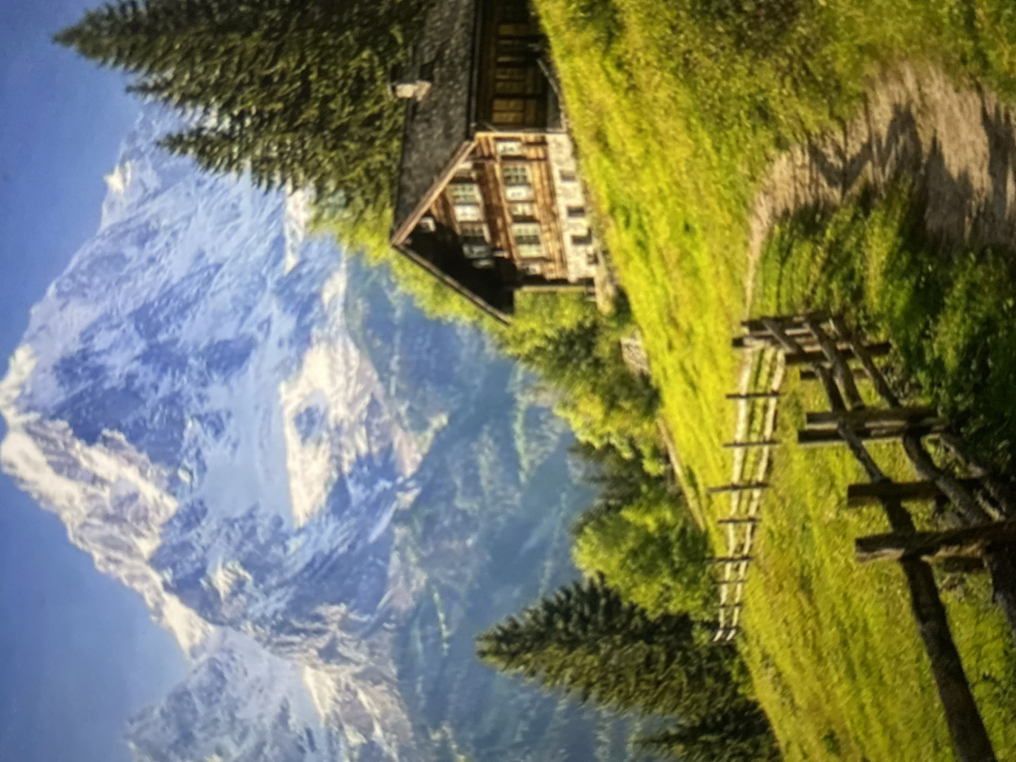 The image depicts a scenic mountain landscape with a wooden cabin situated on a grassy slope. In the background, a majestic snow-capped mountain range dominates the horizon under a clear blue sky. The foreground features a winding dirt path leading up the hill, bordered by a rustic wooden fence. Lush green trees, including tall pine trees and deciduous trees with vibrant green foliage, are scattered across the landscape. The cabin is a traditional alpine style, with dark wood construction and multiple windows. The overall scene evokes a sense of tranquility and natural beauty.
