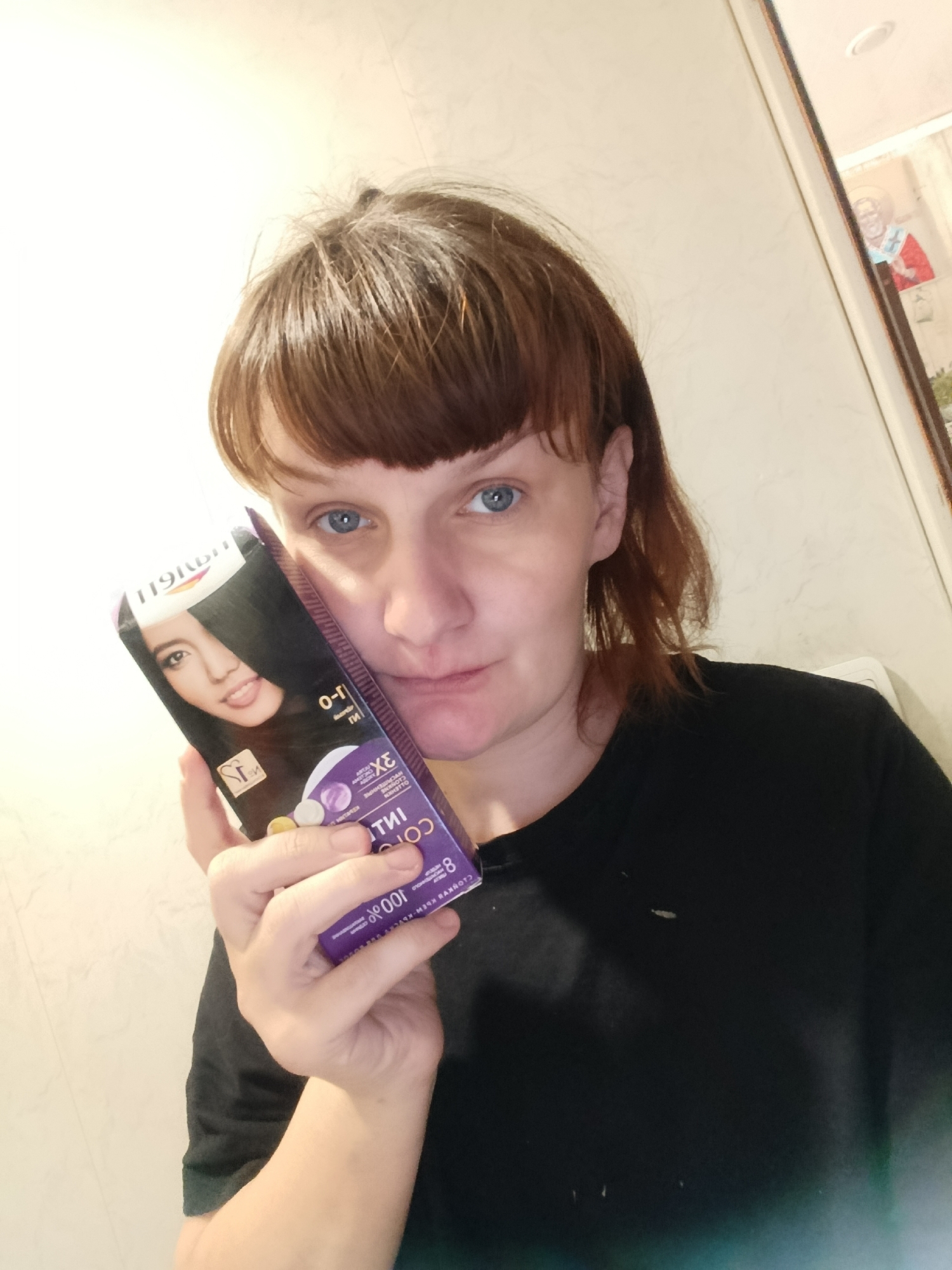 A person with fair skin, blue eyes, and short brown hair is holding a box of hair dye close to their face. The hair dye box is purple and features an image of a woman with dark hair on the front. The person is wearing a black shirt. The background appears to be a bathroom, with a framed picture visible on the wall behind the person. There is no indication of the city of Фоки, Russia, in the image.