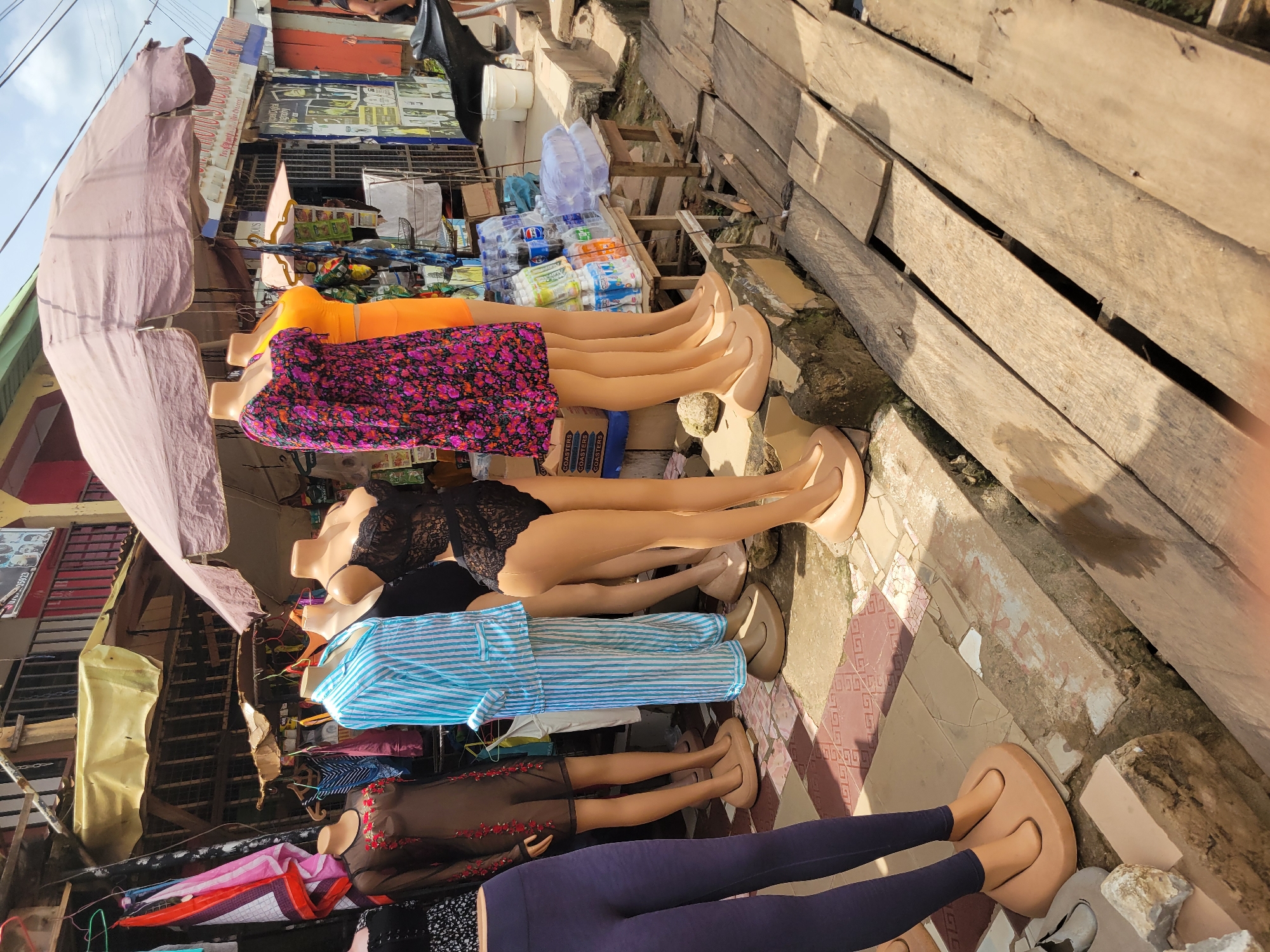 The image shows a market scene in Egbagu, Nigeria. Several headless female mannequins wearing different clothing items (a flower dress, a black lace top, a blue and white striped pajama set, a brown dress with red embroidery, and a navy blue leggings) are displayed on a tiled surface, lined up along a wooden walkway. An awning shades the mannequins and nearby market stalls. In the background, various shopfronts and goods are visible.
