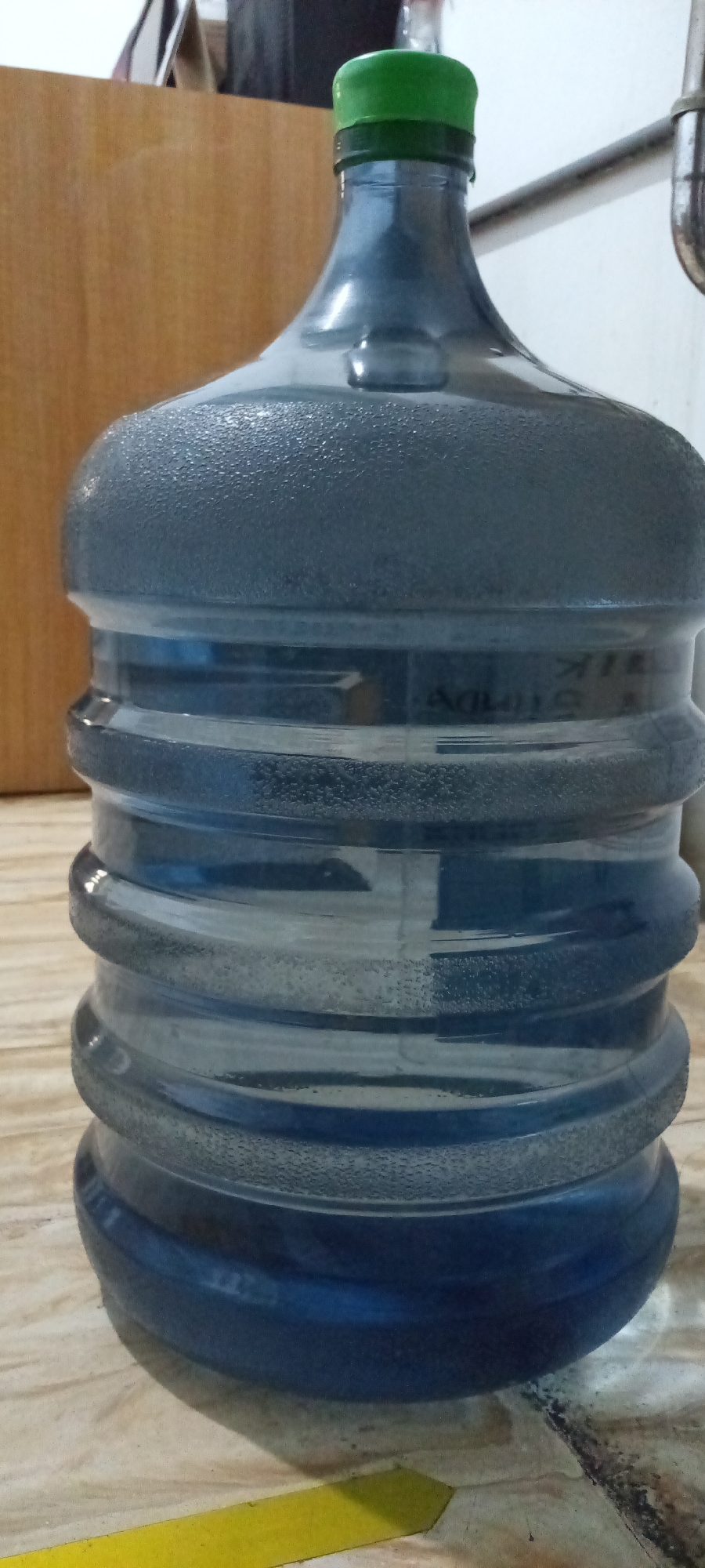 A large, translucent blue plastic water jug with a green cap sits on a tiled floor. The jug is partially filled with water and has a textured surface. In the background, there is a wooden cabinet, a metal pipe, and a white wall. The bottom left corner has a yellow stripe on the floor. The location is implied to be in Lhokseumawe, Indonesia, but no specific landmarks are visible. No people are present in the image.