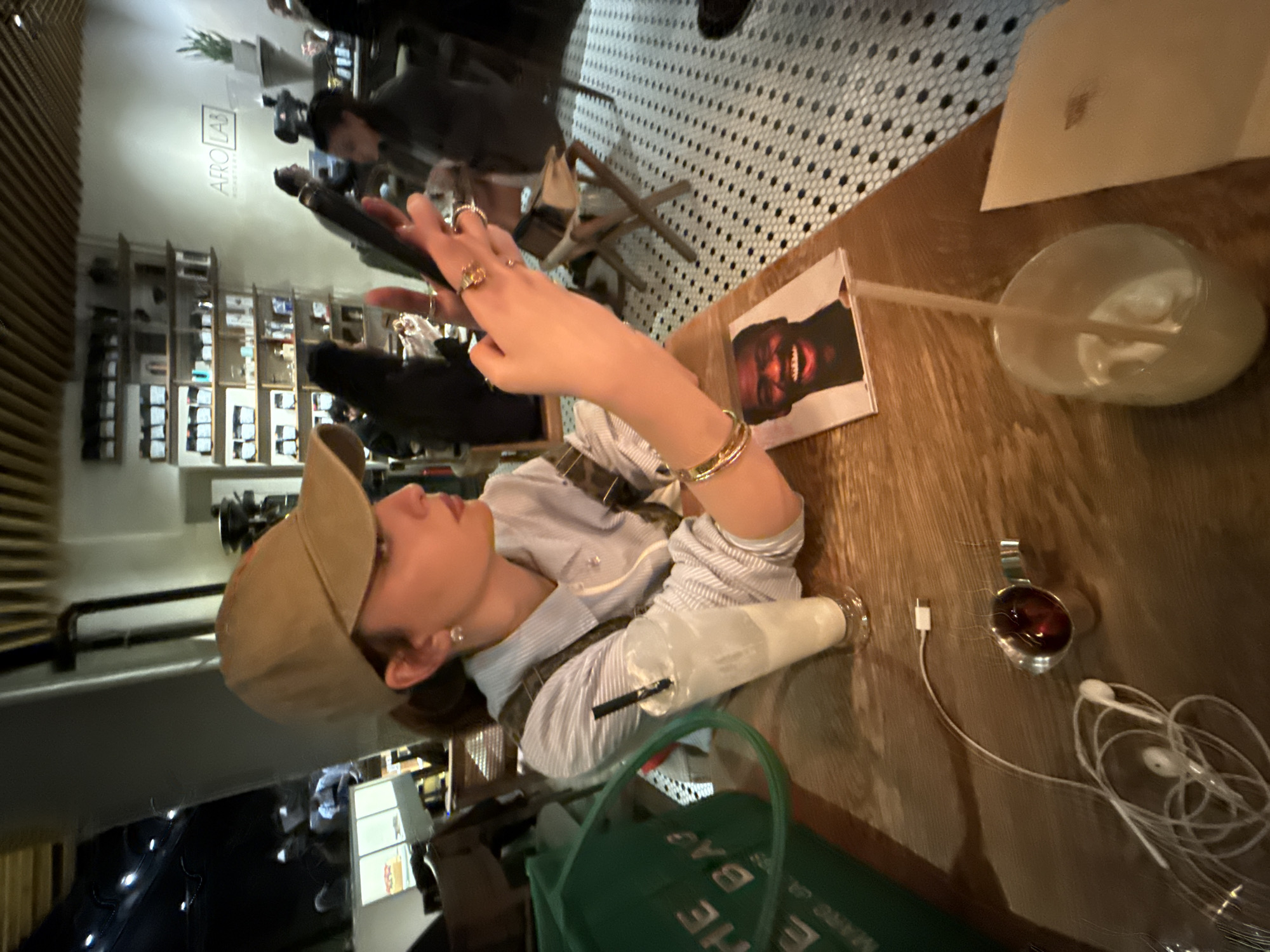 A person wearing a tan baseball cap is seated at a wooden table in a cafe, looking at a phone in their hand. A drink, a small artwork, a charger, and headphones are on the table, along with a green bag. Other patrons are visible in the background, and the cafe has a patterned tile floor and shelves stocked with items. The logo "AFRO LAB ROASTERY" is visible on the wall. Based on the logo and cafe design, this scene likely takes place in Yerevan, Armenia.