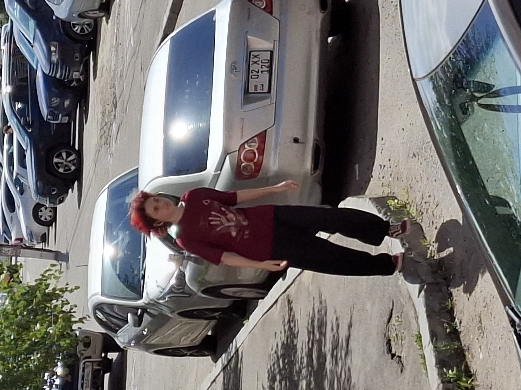 A young person with red hair, wearing a maroon t-shirt and black pants, stands on a curb next to a silver sedan. The person is facing the camera, with their left foot on a small concrete block. Behind the person, a silver sedan is parked, with its rear facing the camera. The license plate of the sedan reads "02 XX 120". To the left of the silver sedan, a white sedan is parked. In the background, several other vehicles are visible, including a dark blue SUV and a lighter colored SUV. The scene appears to be a street or parking lot in a city. The lighting suggests it is daytime, with shadows cast on the ground.