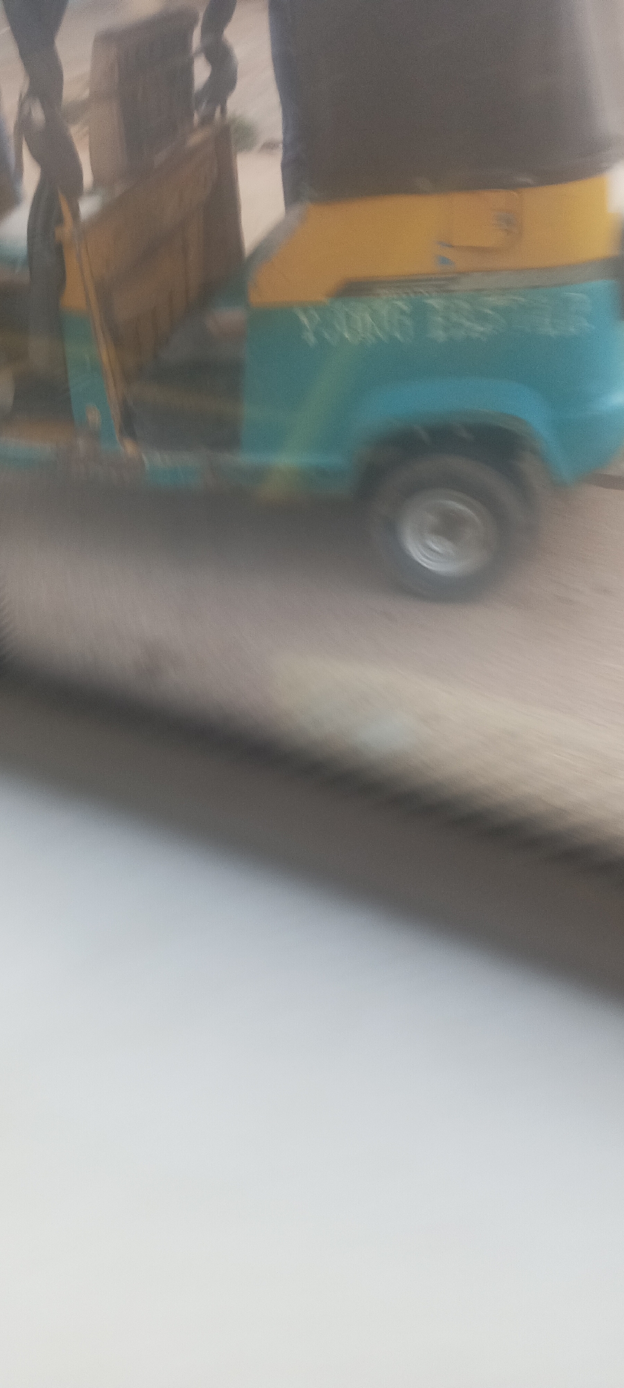 The image captures an outdoor daytime scene, likely taken from a moving or stationary vehicle, looking towards a road. The primary subject is a three-wheeled auto-rickshaw, commonly known as a Keke Napep in Nigeria. The vehicle features a two-tone design, with a teal or light blue body and a bright yellow canopy or upper section.
On the side of the auto-rickshaw, the words "YOUNG BROTHER" are clearly visible in white or light-colored text. A single wheel with a metallic hubcap is visible on the right side of the vehicle. Partially visible figures, likely people, can be seen near the front left side of the auto-rickshaw, suggesting it is either in operation or awaiting passengers.
The road surface appears dusty or unpaved, consistent with outdoor environments in many Nigerian localities, aligning with the provided context of Bukuru. The lighting indicates it is daytime. The lower portion of the image is dominated by a blurred, light-colored surface, possibly the interior of the capturing vehicle or an immediate foreground object. The image has a slight overall blur, which might be due to motion or camera shake, but the details of the auto-rickshaw and text are discernible.