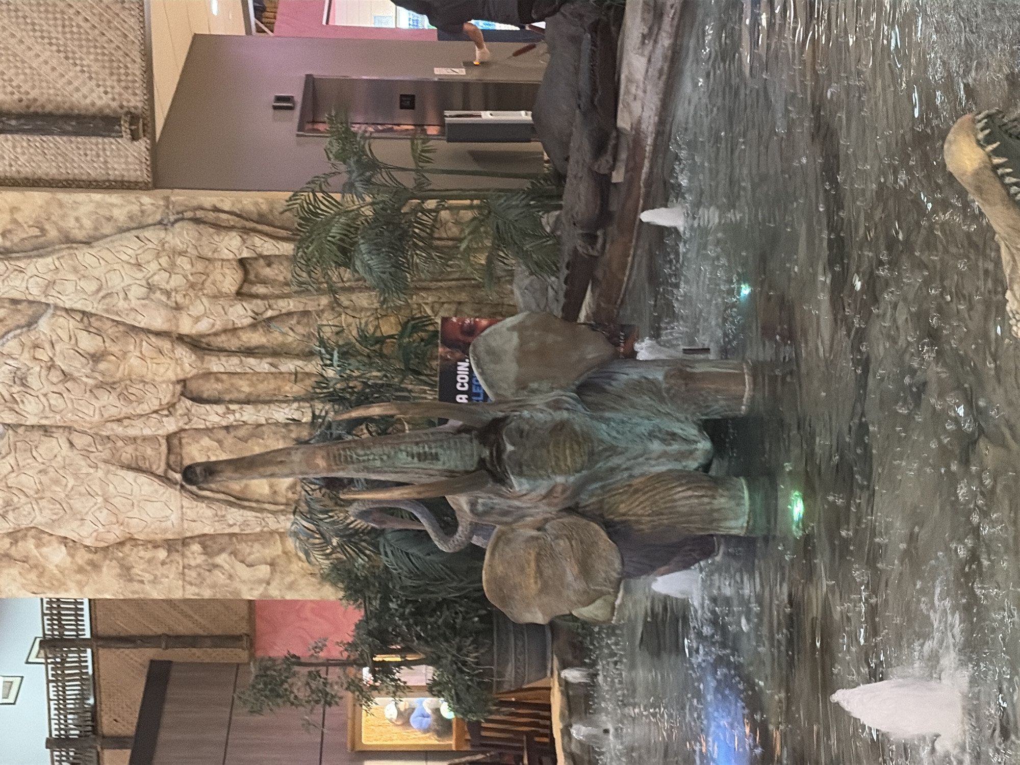 The image depicts an indoor fountain with an elephant statue at its center, surrounded by lush green plants and stone carvings of giraffes on a textured wall. The water in the fountain is disturbed by multiple jets of water, creating a dynamic effect. A partial view of a crocodile sculpture is visible in the foreground, submerged in the water. In the background, an elevator and a woman interacting with an elevator call button can be seen, along with a sign that says "A COIN. ELE". The overall setting appears to be a themed indoor space, possibly a shopping mall or a hotel lobby, with architectural elements suggestive of an African savanna.