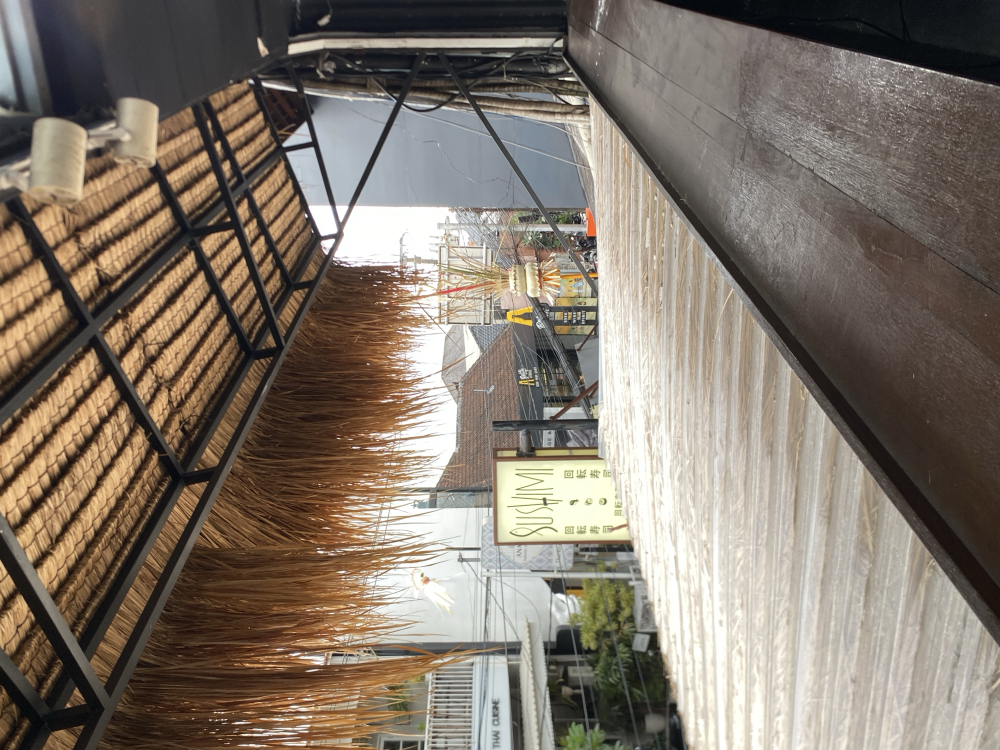An eye-level, medium shot captures an outdoor street view in North Kuta, Indonesia, under a thatched roof structure. The foreground features a textured wooden surface, possibly a table or counter, with a dark brown railing. The background reveals a street scene with buildings, signage, and vegetation. Notably, a sign for "SUSHI" is visible, illuminated with yellow light. The thatched roof extends from the top left corner, its dried fronds hanging down. Overhead, exposed electrical wiring and some white cylindrical objects are suspended. The street appears to be a commercial area, with multiple businesses and shops indicated by their storefronts and signs. The overall impression is one of a casual, outdoor dining or seating area with a view of a bustling street.