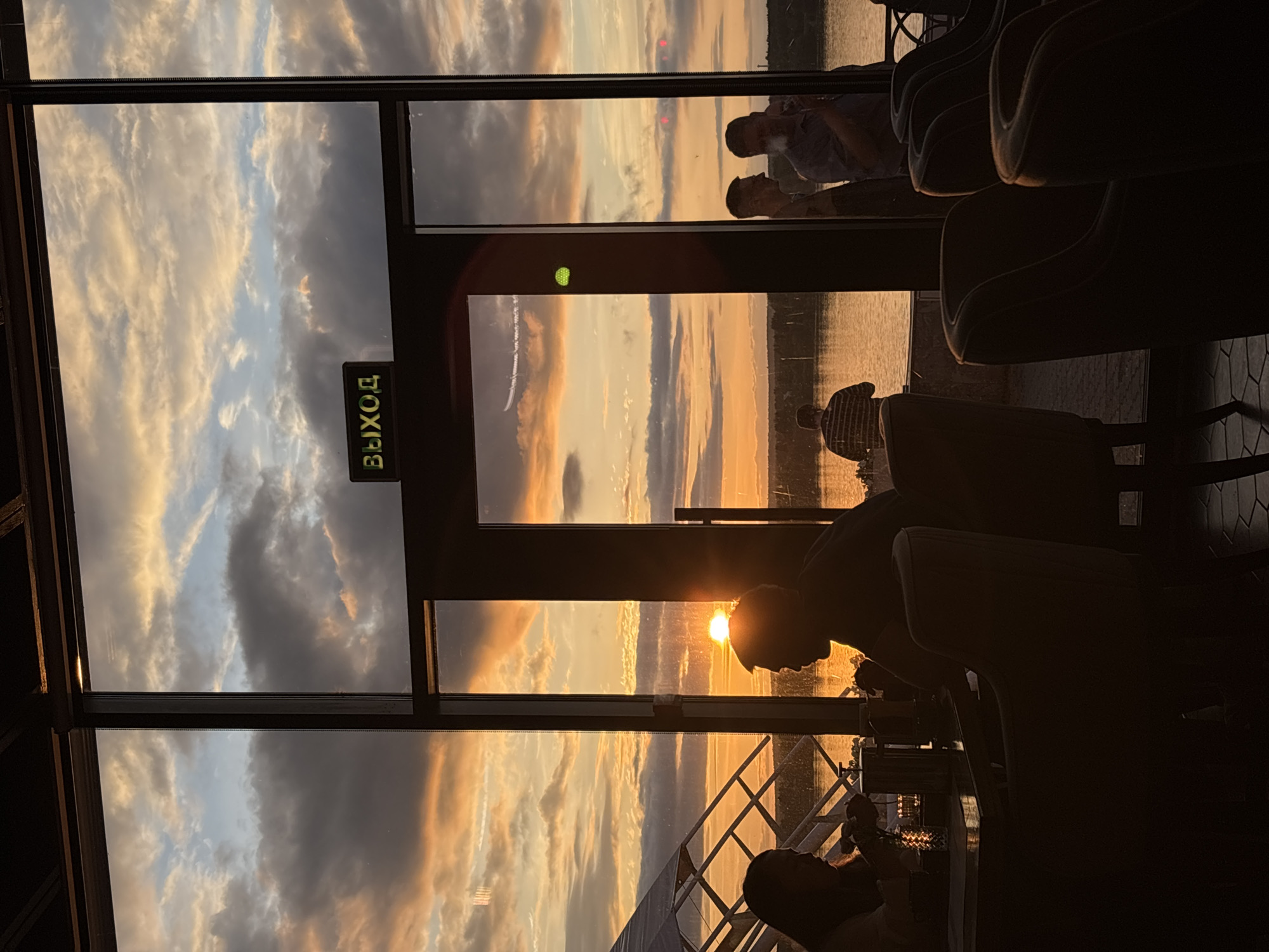 The image captures an interior view of a restaurant or bar at sunset, overlooking a wide river, identified by context as the Kama River in Perm, Russia. The scene is framed by dark-paned glass windows and a central glass door, through which a vibrant sky with orange, yellow, and grey clouds is visible.
In the left foreground, two individuals are seated at a table, their bodies silhouetted against the bright sunset. The person on the far left, with long hair, appears to be looking down at an object on the table, possibly a phone or menu. The person to their right, in profile, is facing left. A small, illuminated decorative light is on their table. Several dark, upholstered armchairs are positioned in the immediate foreground, with hexagonal patterned floor tiles visible beneath.
Above the central glass door, a black rectangular sign with illuminated green Russian text "ВЫХОД" (EXIT) is prominently displayed.
Through the large glass panels, the sun is visible as a bright disc near the horizon, casting an intense orange glow across the water. A light-colored, geometric structural framework, possibly part of a canopy or building extension, is visible on the far left of the exterior. A person wearing a striped shirt and a cap is standing by the water's edge, facing away from the camera, on the left side of the exterior view. On the right side, two men are standing outside or in a reflection within the glass, one of whom is exhaling a plume of smoke or vapor. A faint white contrail extends across the sky through the rightmost glass panel. A small green circular light is affixed to the right glass panel, with distant red lights also discernible. The interior of the establishment is largely dark, silhouetting the occupants and furniture against the dramatic sunset.