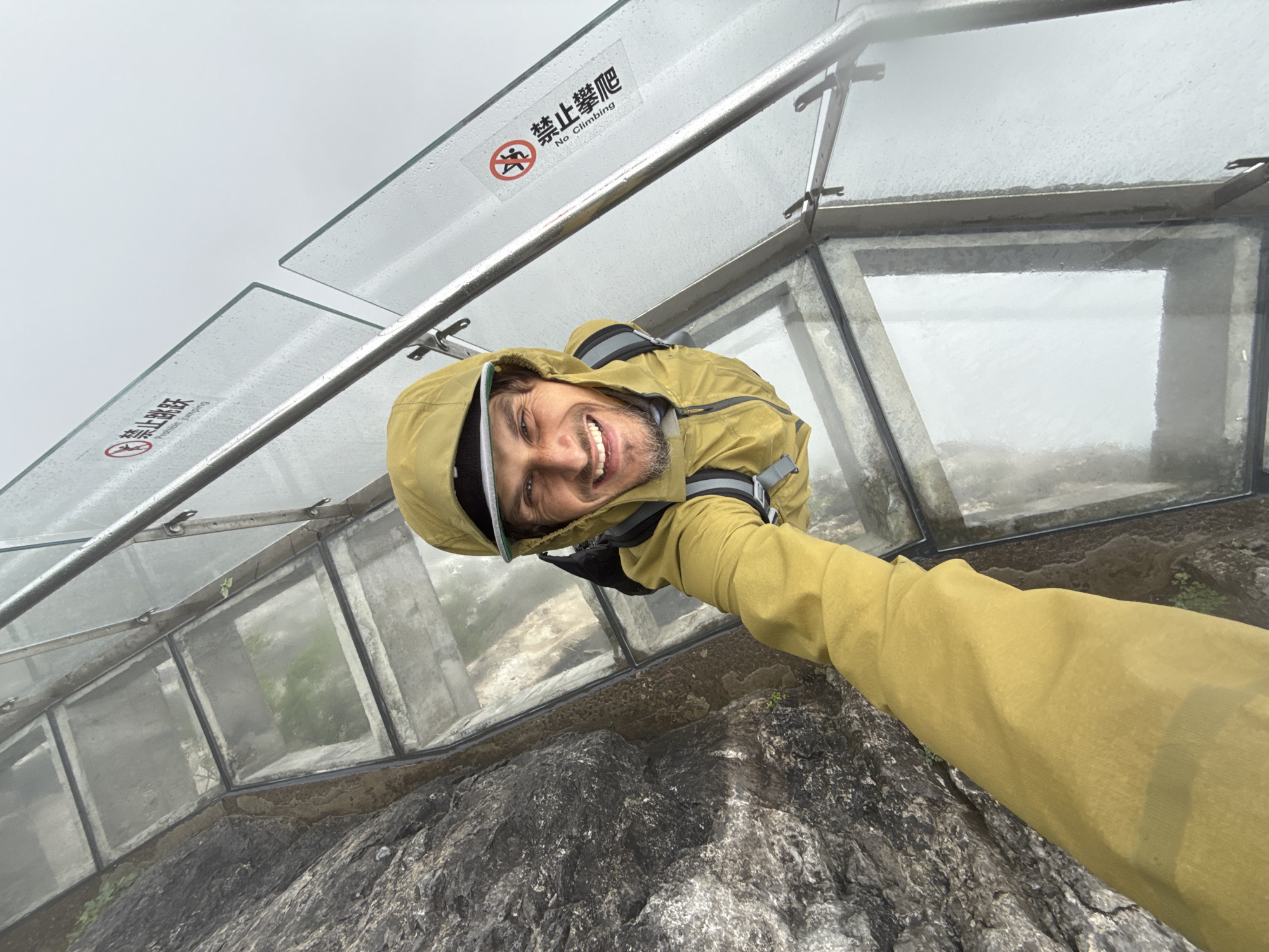 The image shows a man in a yellow jacket and hood taking a selfie while standing on or near a glass-bottomed walkway. He is smiling and looking at the camera. The walkway has a metal railing and a sign in Chinese and English that reads "No Climbing." The walkway appears to be built on a rocky terrain. The image suggests a high-altitude location, likely in a mountainous area with foggy or cloudy weather. Based on the sign and context clues, the location is possibly in the Yongding District, China.