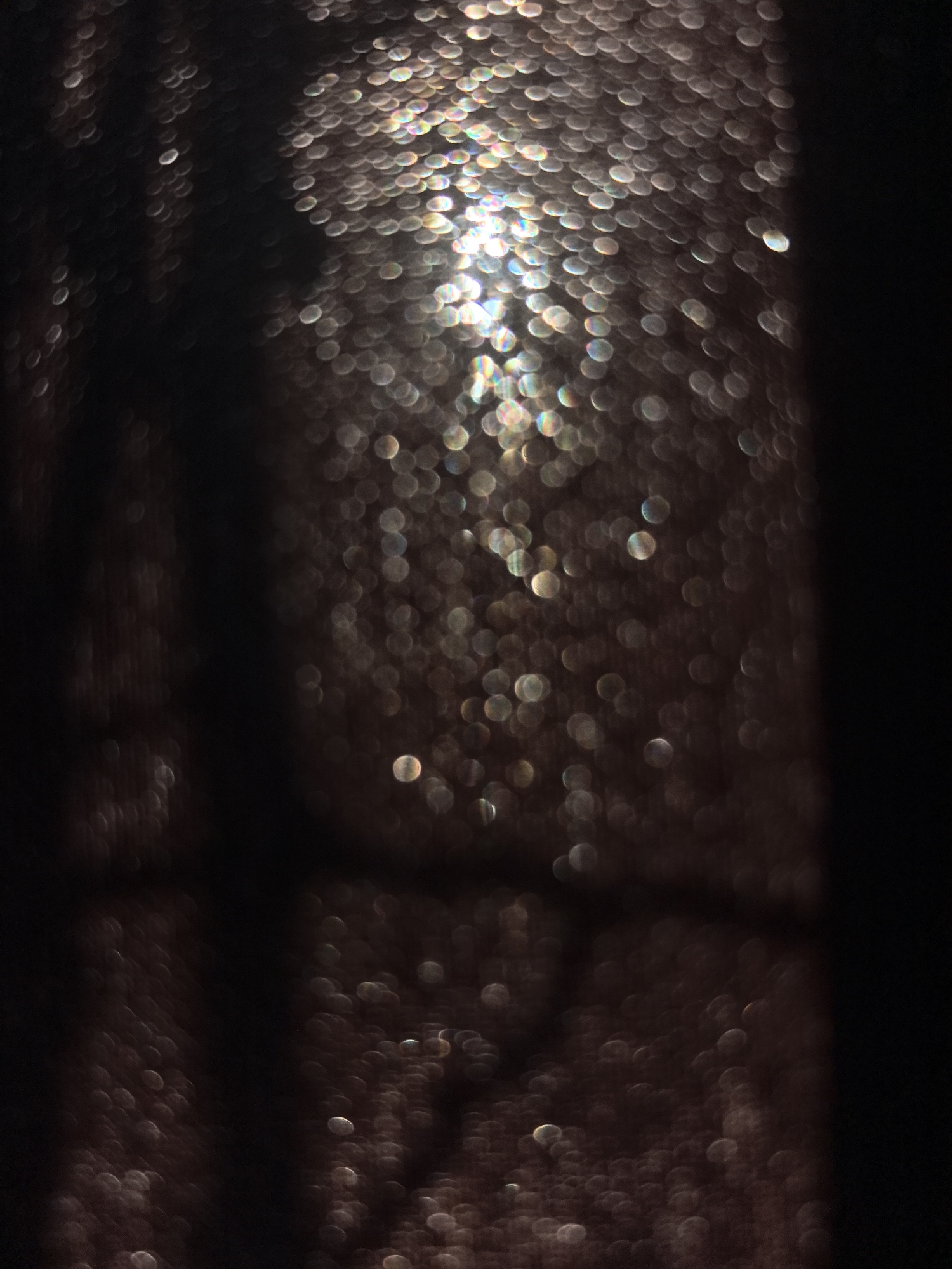 The image shows an out-of-focus view through a window covered in water droplets. The water droplets create bokeh, reflecting a bright light source in the center, surrounded by a dark environment. The details are obscured. The location is not discernible.