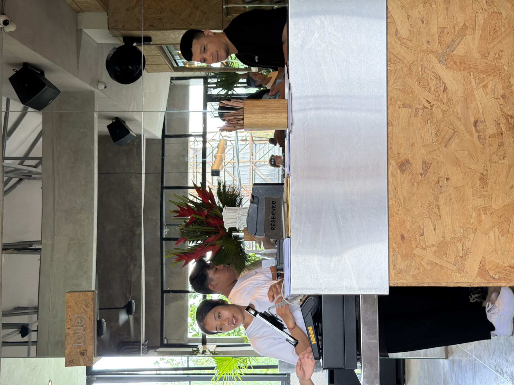 Captured indoors, the image shows two individuals standing at a counter. One person, dressed in a white shirt, faces the camera directly. The second person, wearing a black shirt, faces away. Various objects are arranged on the counter, including a vase of flowers, a "RESERVED" sign, and utensils. A sign reading "toilet" is visible on the left. The environment includes an outdoor view, visible through windows, which suggests an urban setting. Based on the information, this scene is likely located in North Kuta, Indonesia.