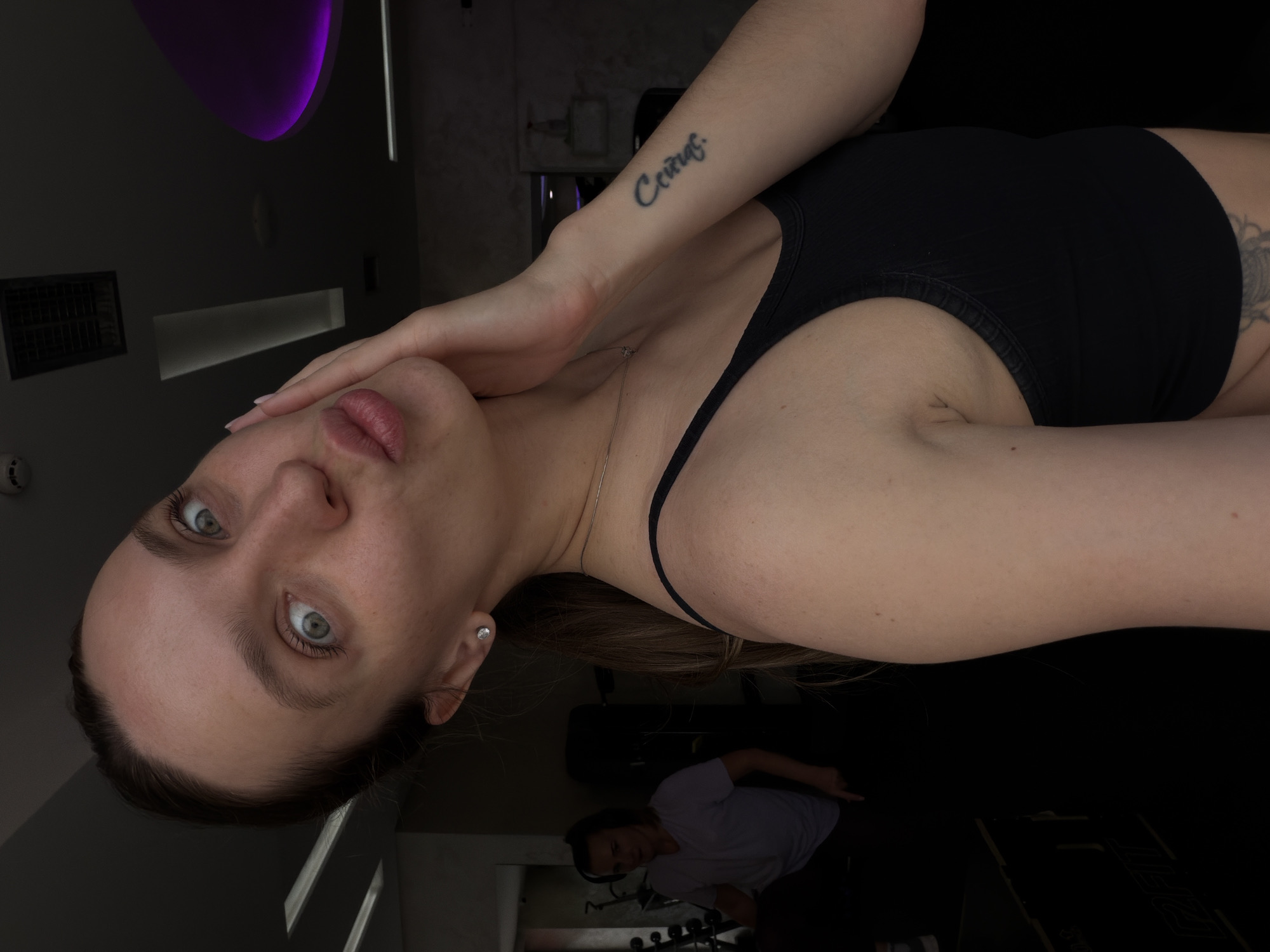 A medium, canted, and somewhat dimly-lit indoor shot depicts a young woman in a black top with fair skin, facing the camera. She has light eyes and pursed lips. Her right hand is placed near her face. On her left forearm, the word "Сейчас" is tattooed. A silver chain is visible around her neck.
In the background, a person is vaguely visible sitting on a bench, and there is exercise equipment. A purple light fixture can be seen on the ceiling. The location may be a gym or fitness studio.
It is impossible to confirm the location as Budva, Montenegro, based on the image alone.