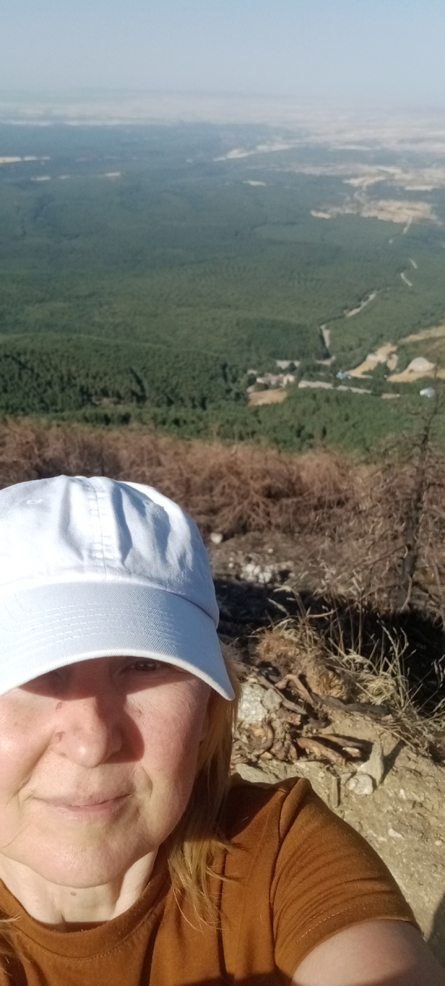 A selfie is taken outdoors, capturing a woman wearing a white baseball cap and an orange t-shirt. The woman is positioned in the foreground on the right side of the frame, with her face partially visible. Her eyes are looking towards the camera, and she is smiling. Behind her, the landscape unfolds with a vast expanse of green pine forest. In the distance, a hazy horizon suggests more undeveloped land or a body of water. The foreground shows dry, brown brush and some rocks, indicating a natural, possibly arid, environment. The image appears to be taken from a high vantage point, possibly a hilltop, offering a panoramic view of the surrounding area. The lighting suggests it is daytime, with sunlight casting shadows. The location is identified as being in Cerezo de Arriba, Spain.