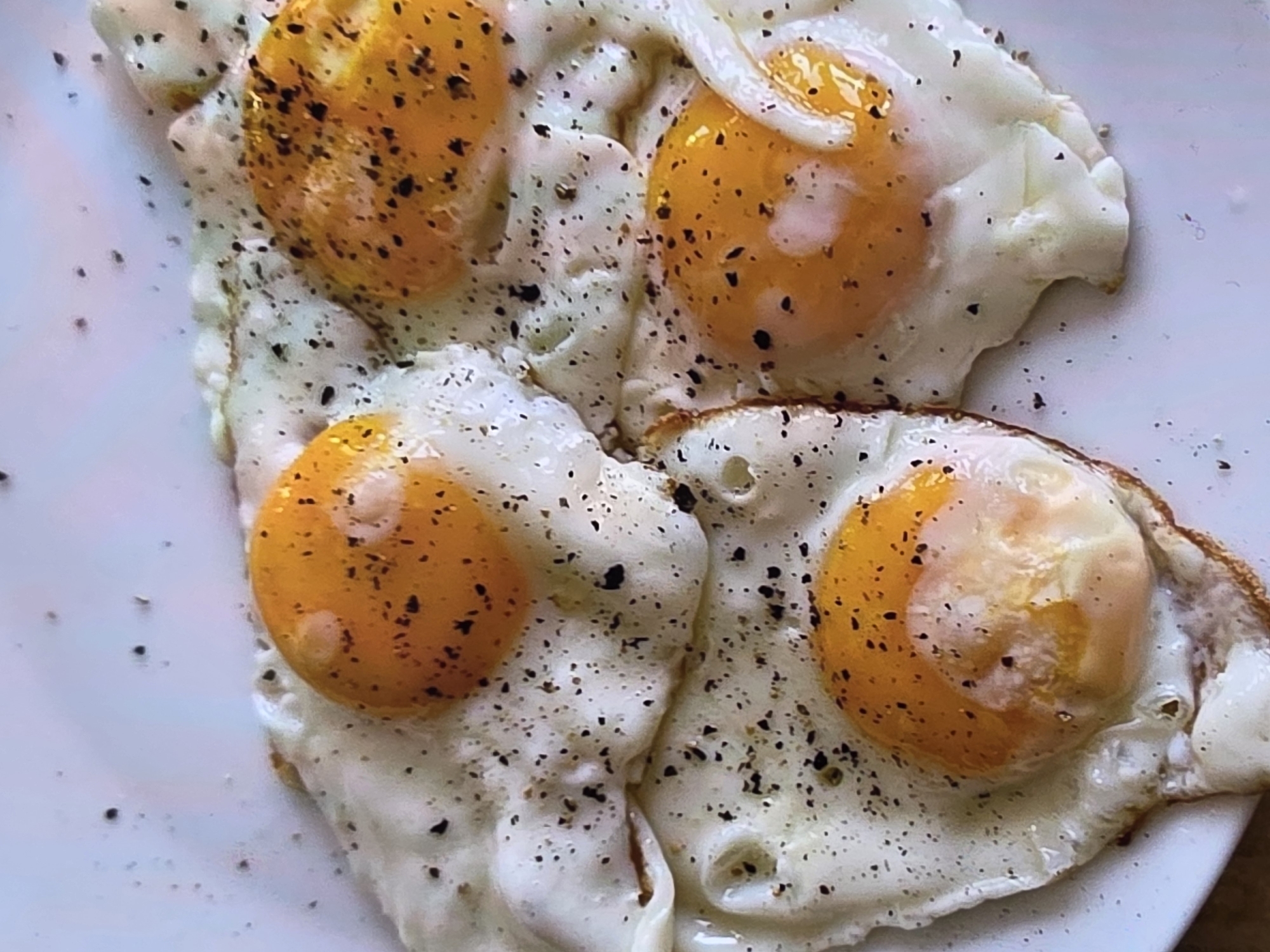 The image displays four fried eggs seasoned with black pepper, arranged on a white plate. The eggs are sunny-side up, with visible yolks and cooked whites. The background is a plain white surface. There is no discernible indication of the location in the city of Voluntari, Romania.