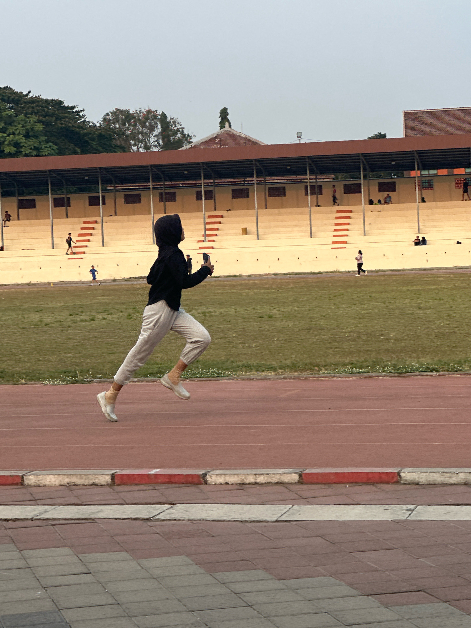 In Mojokerto, Indonesia, a young woman in a black hijab and light grey athletic wear is captured in mid-stride on an outdoor running track. She is actively engaged in running, holding a smartphone in her right hand. The track is a reddish-brown color, with a white line marking its edge. Beyond the track, a grassy field stretches out, and further back, a tiered stadium seating area is visible. Several other individuals are scattered throughout the stadium, some sitting and others walking. The sky above is overcast, suggesting an ambient light condition. The overall scene depicts a public athletic facility with people participating in or observing sports activities.