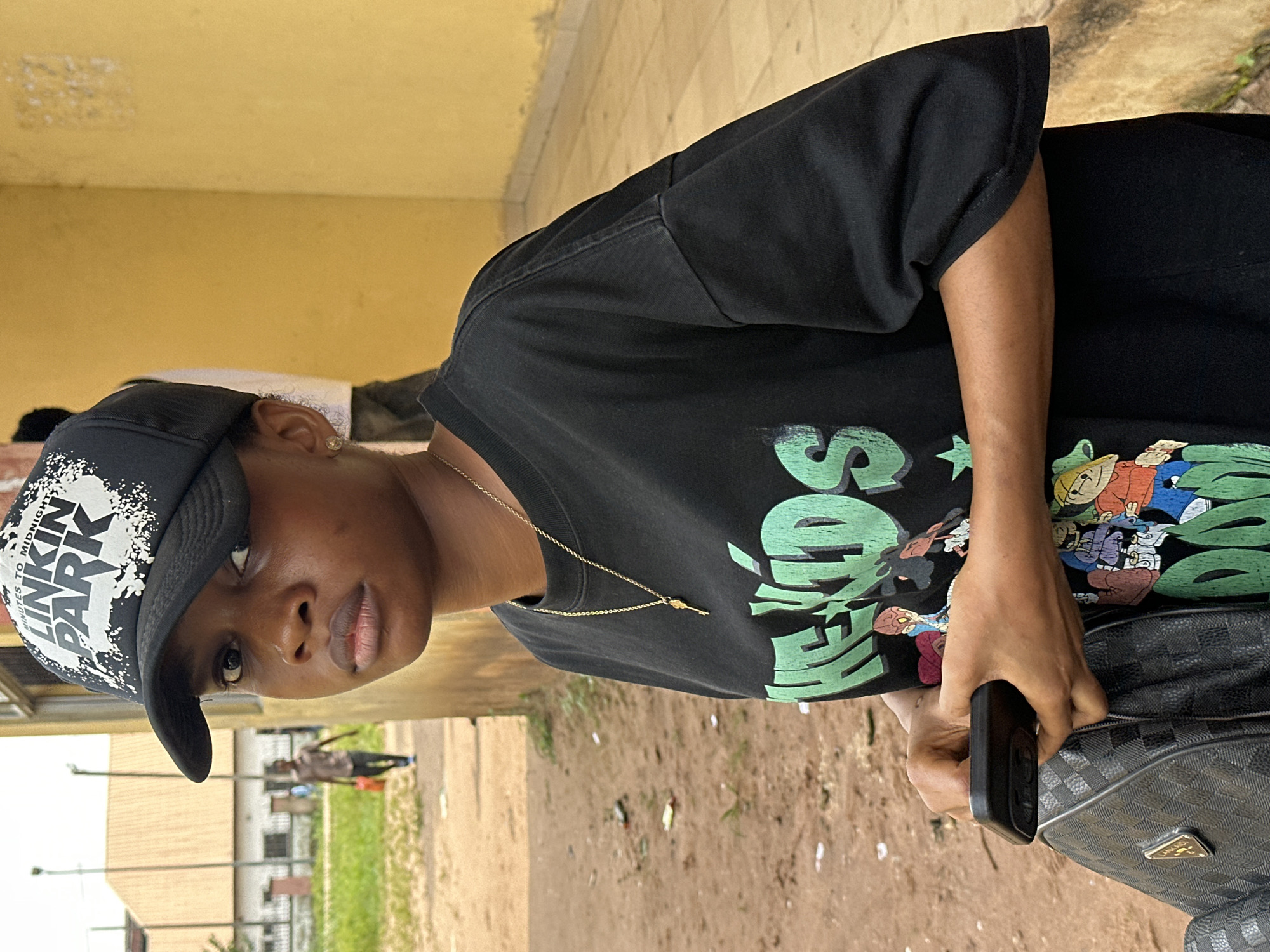 A medium shot shows a young Black woman wearing a black baseball cap with the text "LINKIN PARK" and a black t-shirt with a green graphic that appears to read "HEAPS" with cartoon characters. She is wearing a gold necklace with a small key pendant. She is holding a black smartphone in her left hand and a black backpack is visible on her left side. The woman is standing in front of a yellow building with a rough texture. The ground is dirt and gravel. In the background, a person in a brown shirt and pants is walking away from the camera, carrying an orange bag. The environment appears to be an outdoor, urban setting, possibly in Ihiagwa, Nigeria, given the context.
