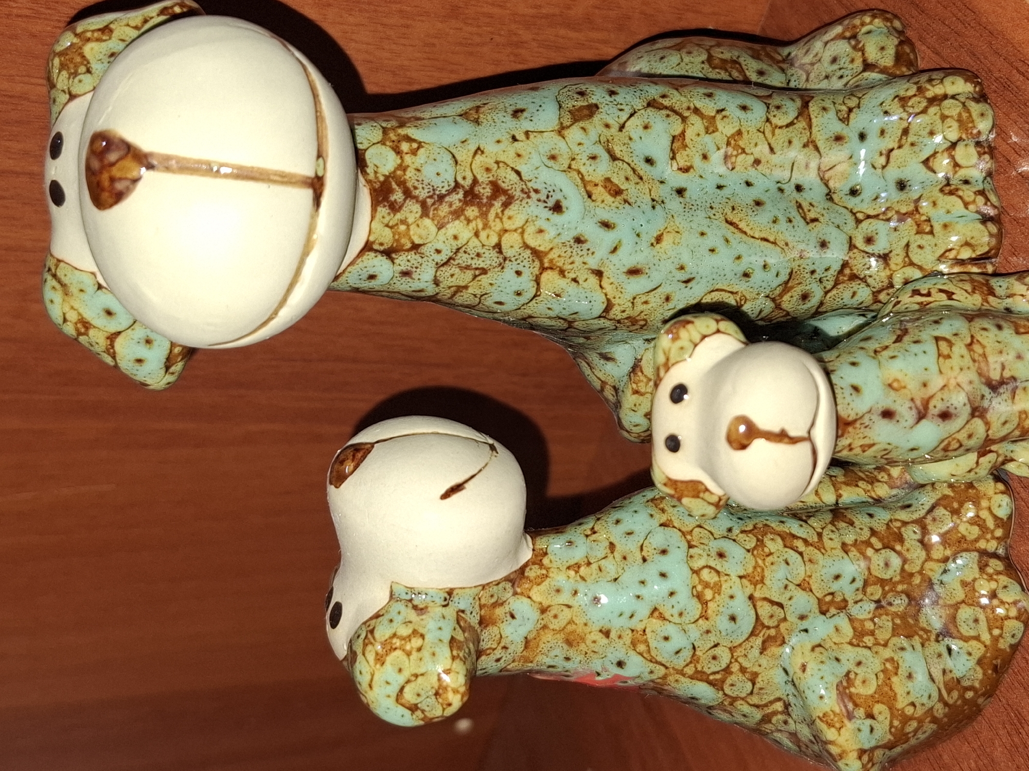 Three ceramic dog figurines are displayed on a wooden shelf. The figurines have a speckled glaze in turquoise and brown. The largest figurine is in the center, with a smaller figurine to its left, and the smallest to its right. The background is a plain wooden surface. The image was taken indoors. The specific location in Almaty, Kazakhstan, cannot be determined from the image.