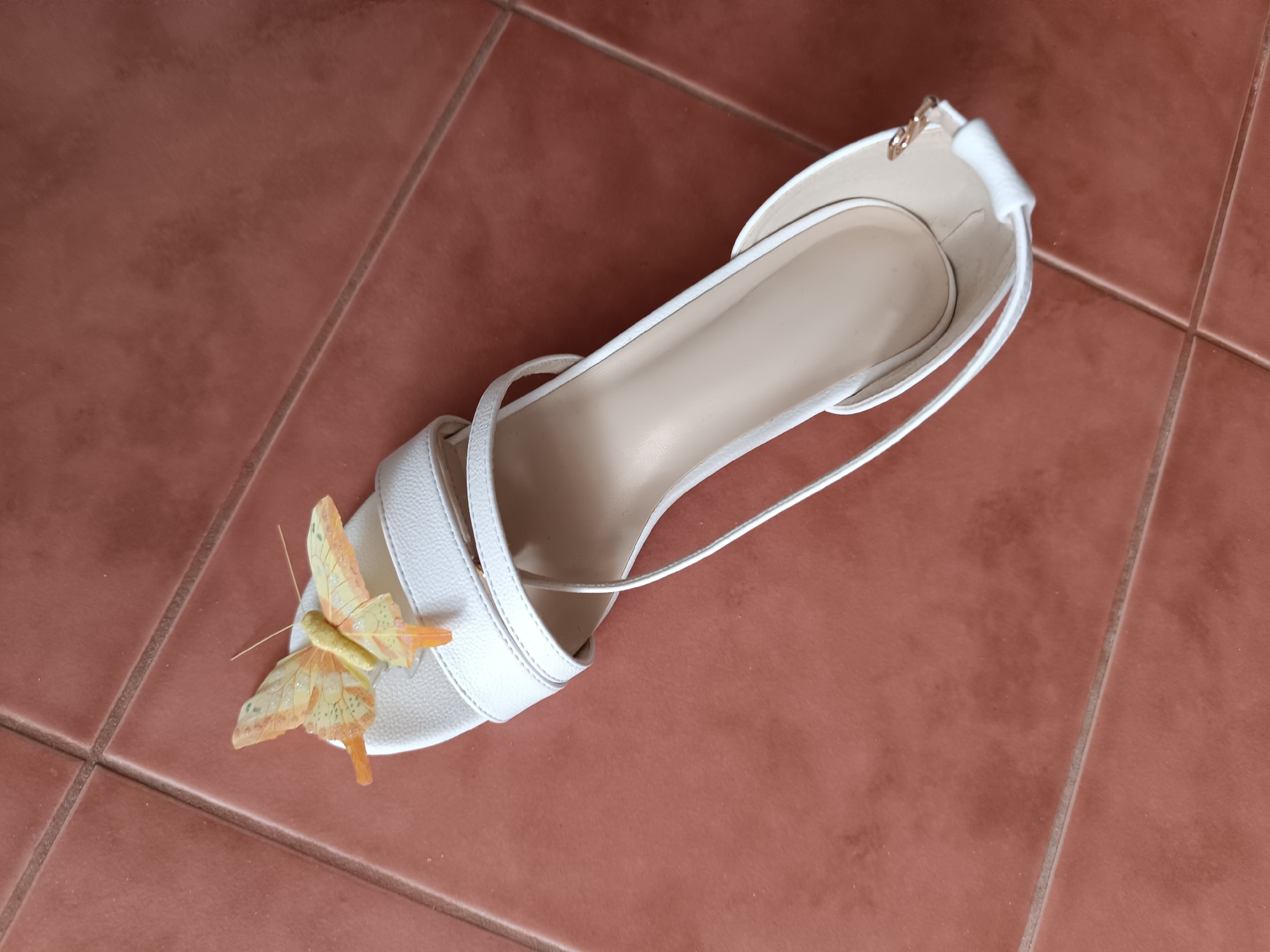 A white stiletto heel with a butterfly decoration is lying on terracotta-colored tiles. The shoe has thin straps and a pointed toe. The butterfly is yellow and orange with glitter accents and is positioned on the front of the shoe. The background consists of a tiled floor. The image appears to be taken indoors. There is no indication of the location being Ternopil, Ukraine.