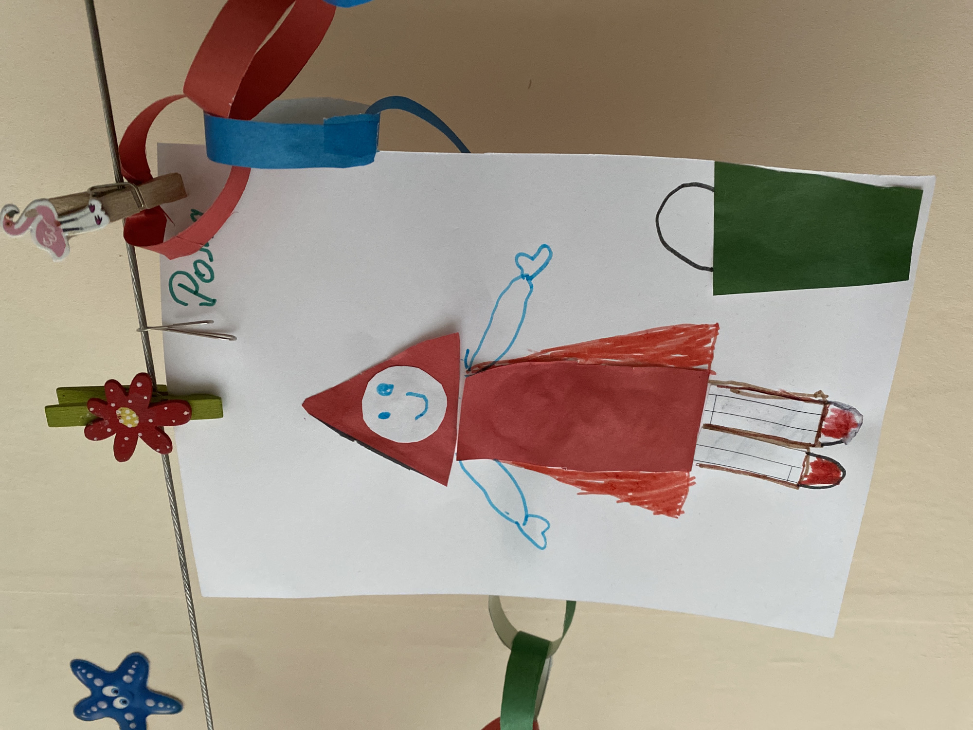 The image shows a child's drawing of a rocket or superhero figure attached to a display. The figure has a red triangular head with a simplistic face drawn on it. The body is constructed from red paper and crayon. Blue lines are used to depict arms with hands shaped like hearts. Beneath the figure are two crayon drawings of what may be rockets with red tips.
The drawing is hung with a series of clips and paper chain decorations, including a flamingo-shaped clip, a flower clip, and a blue starfish clip. The background is a plain beige wall.
There is no information in the image to suggest its location is Yekaterinburg, Russia.