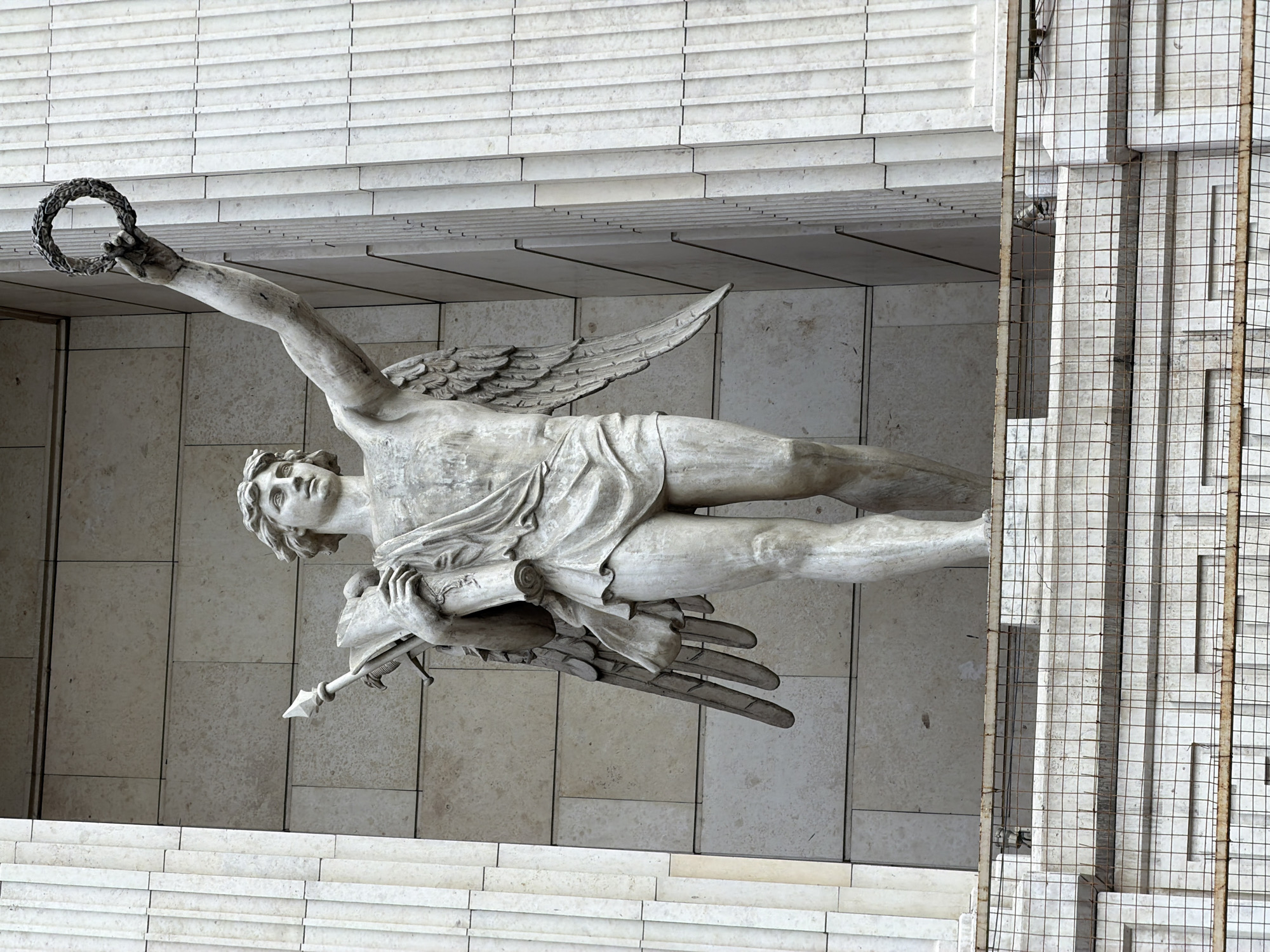 The image shows a stone statue of a winged figure holding a wreath in its raised right hand and a scroll and spear in its left arm. The statue is positioned within an alcove supported by fluted columns. The figure is depicted in a dynamic pose, with its left leg forward, suggesting movement. The statue is made of weathered white stone, with visible detailing on the wings, drapery, and facial features. The background consists of a tiled wall and architectural elements of a building. In the foreground, there is a wire mesh barrier and architectural detailing. The statue is identified as the Victory monument on Nevsky Prospekt in Saint Petersburg, Russia.