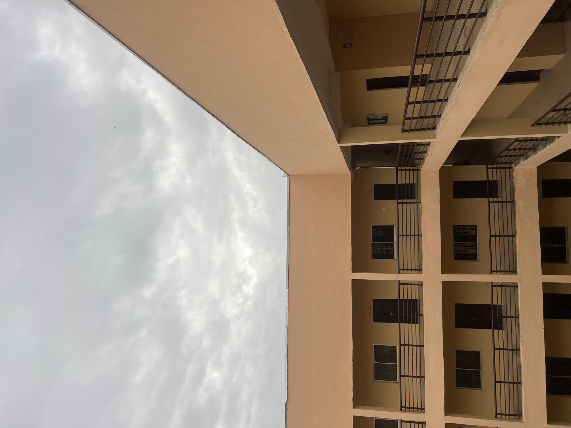 This is a photograph of a beige, multi-story building under a cloudy sky in Wumat, Nigeria. The building appears to be a dormitory or apartment complex, with multiple windows and doors visible on each level. A dark metal railing runs along the balconies on each floor, providing a barrier. The overall impression is one of a modern, but somewhat stark, architectural structure. The sky is overcast with gray clouds, indicating a potentially humid or rainy day. There are no people or vehicles visible in the image. The scene is captured from a slightly elevated angle, looking upwards towards the building and sky.