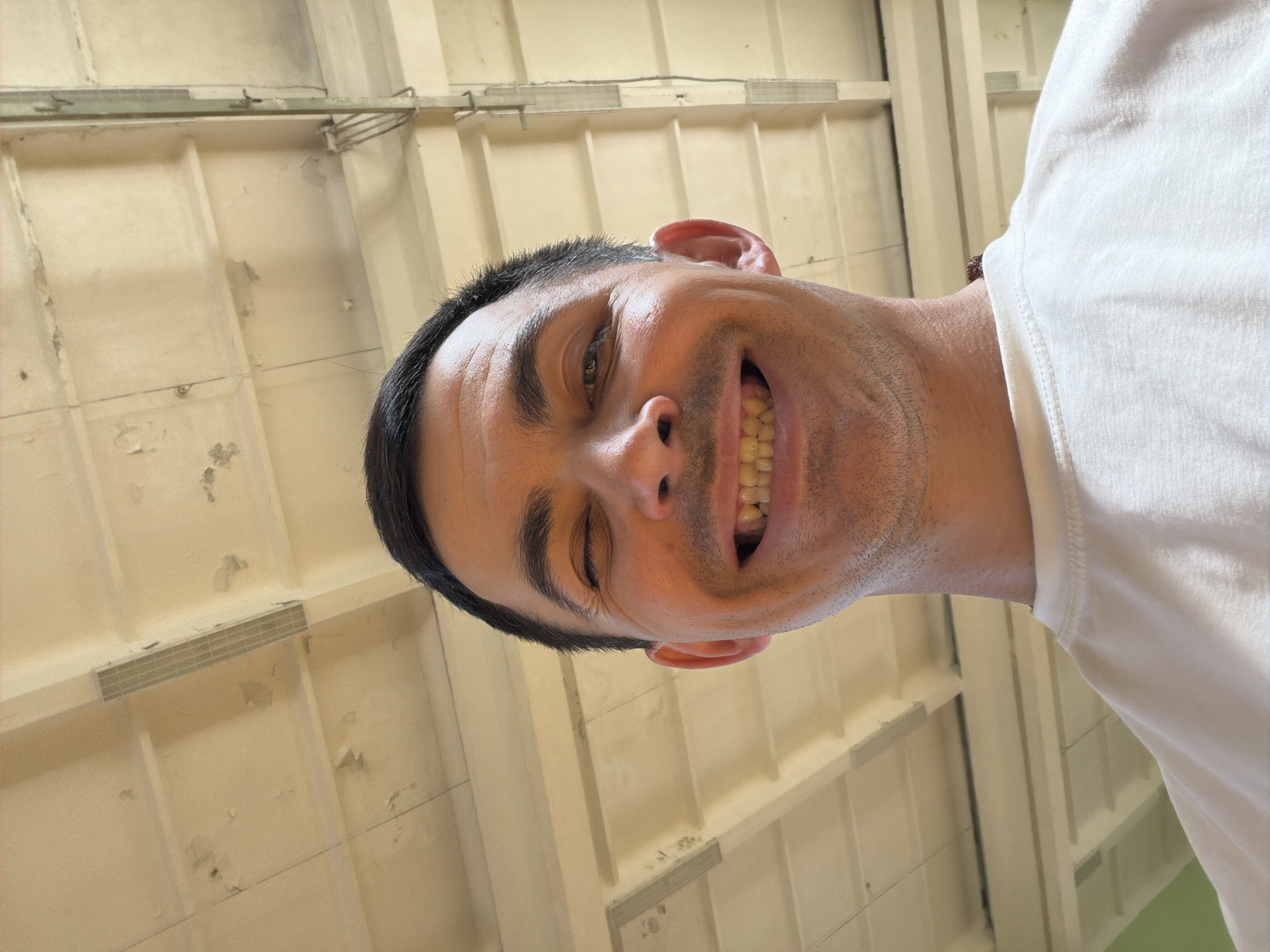 A man smiles broadly while standing in an interior space with a ceiling composed of pale yellow panels. He is wearing a white t-shirt. The location is not provided in the image or the request.