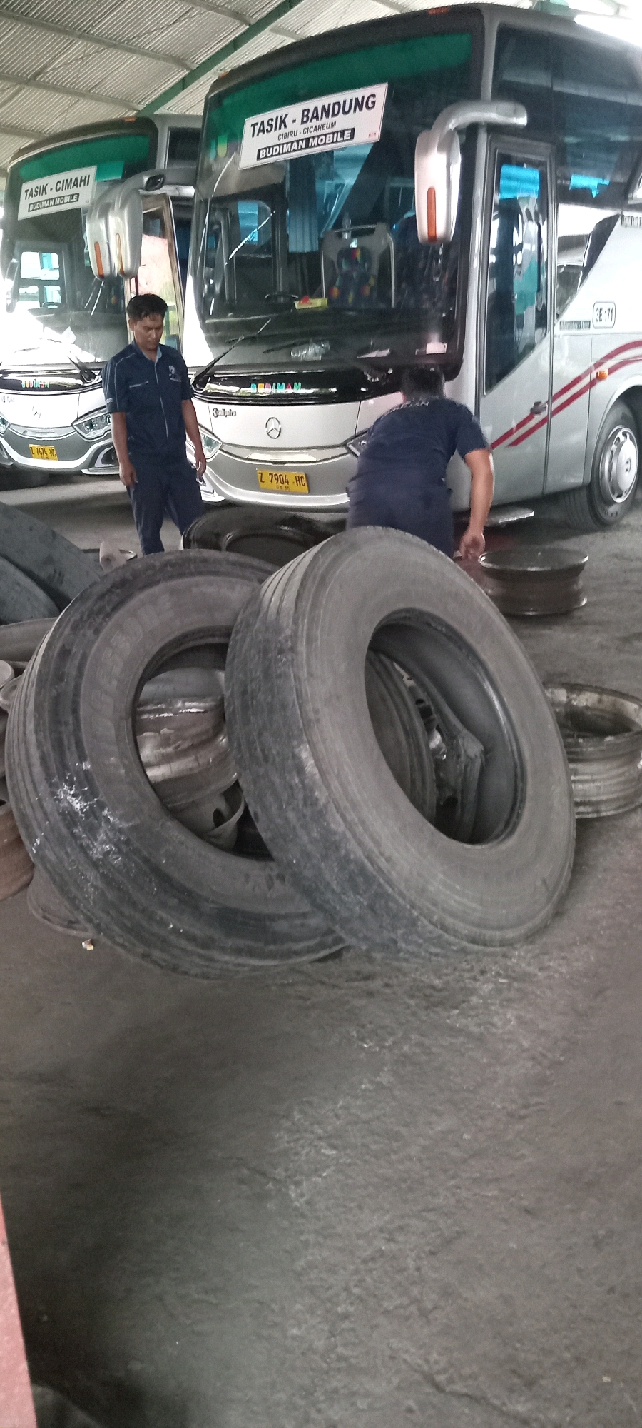 In an indoor garage or maintenance facility in Tasikmalaya, Indonesia, two men are visible. One man, wearing a dark blue uniform with a collar, stands in the center of the frame, looking towards a bus. He appears to be a mechanic or employee of the bus company. Another man, also in a dark blue uniform, is seen from behind on the right side of the image, bent over near the wheels of a bus.
In the foreground, several large, used tires are piled up, some stacked on top of each other, suggesting tire maintenance or storage. The tires are dark in color and appear worn.
Behind the tires, two buses are parked. The bus on the left has a sign that reads "TASIK - CIMAHI BUDIMAN MOBILE". The bus on the right has a sign that reads "TASIK - BANDUNG CIBIRU - CICAHEUM BUDIMAN MOBILE" and a license plate reading "Z 7904 HC". The buses are modern passenger coaches, silver and white with red stripes. The environment suggests a bus depot or repair shop, indicated by the presence of the buses, tires, and the utilitarian concrete floor and ceiling structure. The lighting appears to be artificial, consistent with an indoor space.