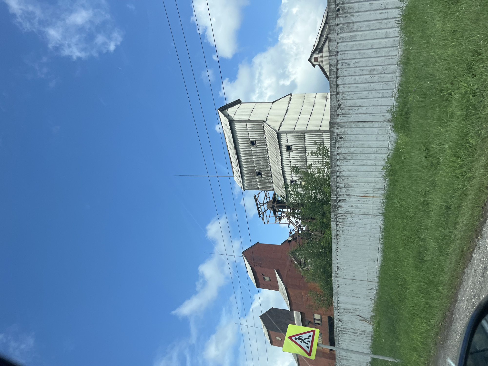 A street-level, eye-level, medium shot captures an industrial area under a bright blue sky with scattered white clouds. In the foreground, a bright yellow pedestrian crossing sign with a red border and a black silhouette of a person walking is prominently displayed on a pole to the left. A grey, corrugated metal fence runs horizontally across the middle of the frame, separating the foreground from the industrial structures behind it. Tall green grass grows in front of the fence.
Behind the fence, several tall, multi-story industrial buildings are visible. The building on the far left is a dark red structure with a peaked roof. Adjacent to it is another dark red building, slightly taller and with a more weathered appearance. To the right of these, a partially constructed or disused metal frame structure rises. Further to the right, a tall, white, corrugated metal building with a peaked roof and several small, dark rectangular openings is the most prominent structure. To its right, a lower, white, corrugated metal building with a single window is visible. The overall impression is one of an active or former industrial zone, likely located in or near the city of Серково, Russia. Overhead, dark power lines stretch across the sky.