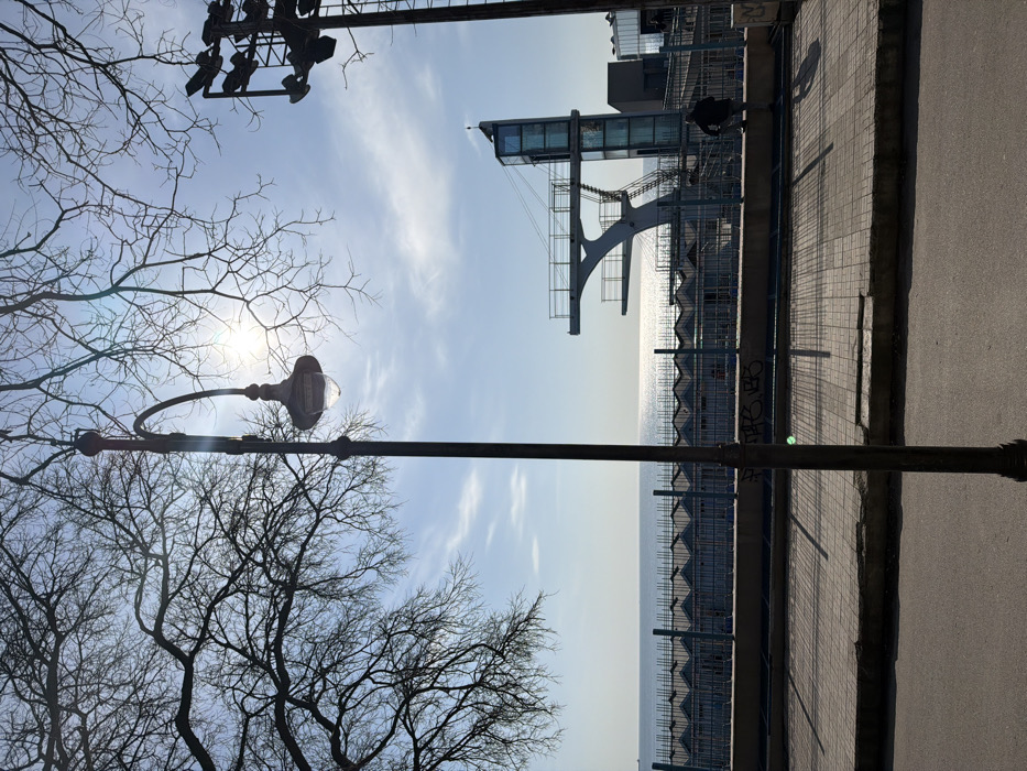 This image depicts a coastal urban scene on a clear, bright day in the city of Unknown, Unknown. In the foreground, an asphalt road runs along the bottom, leading to a light-colored paved sidewalk with visible cracks. A dark, ornate streetlamp post stands centrally, extending from the bottom, featuring an upward-curving arm holding a clear, bell-shaped lamp. Graffiti is present on a low concrete barrier separating the sidewalk from a lower area.
Beyond this barrier, a blue metal fence runs horizontally. Behind the fence, a long row of uniform, light-colored beach huts or changing cabins with gabled roofs stretches towards the horizon, facing the sea. To the right, a prominent multi-level diving platform structure, primarily white or light grey, features railings, stairs, and a clear or blue glass-enclosed tower or elevator shaft. A single individual, dressed in dark clothing, is observed standing in silhouette on a lower platform near the base of the diving structure, facing towards the right. Partially visible on the far right is a tall metal structure equipped with multiple floodlights.
The background features a calm, expansive blue sea with significant sun glare reflecting off the water's surface. Above the horizon, the sky is light blue with scattered wispy clouds. Bare, dark tree branches frame the upper portions of the image, silhouetted against the bright sky and the visible sun, which causes a lens flare near the top center.