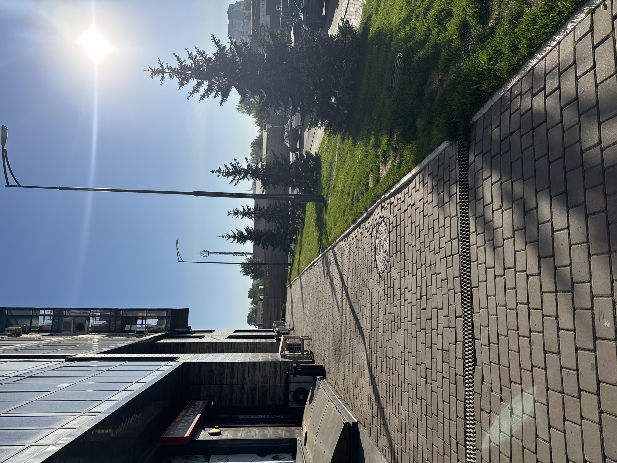 A brightly lit urban street scene in Krasnoyarsk, Russia, unfolds on a clear, sunny day. The image shows a wide, paved pedestrian sidewalk on the right, constructed from light gray rectangular pavers. A metal drainage channel runs parallel to the curb, which separates the sidewalk from a strip of well-maintained green grass. Along this grassy strip, several coniferous trees (likely firs or spruces) cast long shadows onto the sidewalk, indicating a mid-day sun position.
Beyond the trees, a road is visible with several parked cars, including a white and red sedan. In the background, further urban buildings and structures can be seen. On the left side of the frame, a modern, multi-story building with dark, reflective glass panels and dark textured siding dominates the foreground. Numerous outdoor air conditioning units are mounted on its exterior.
Two streetlights with long, horizontal arms extend into the clear blue sky. The sun is prominently featured in the upper left, casting a strong glare and bright light across the scene.
Visible Cyrillic text on a red sign attached to the building on the left reads "ПРОДУКТОВЫЙ" (PRODUKTOVY), indicating a grocery or food store. In the distant background, partially obscured signs contain fragments like "АВТО" (AVTO - Auto) and "ГРУЗОВОЙ" (GRUZOVAY - Cargo/Truck), suggesting automotive-related businesses. The overall scene depicts a typical, clean, and well-maintained city environment.