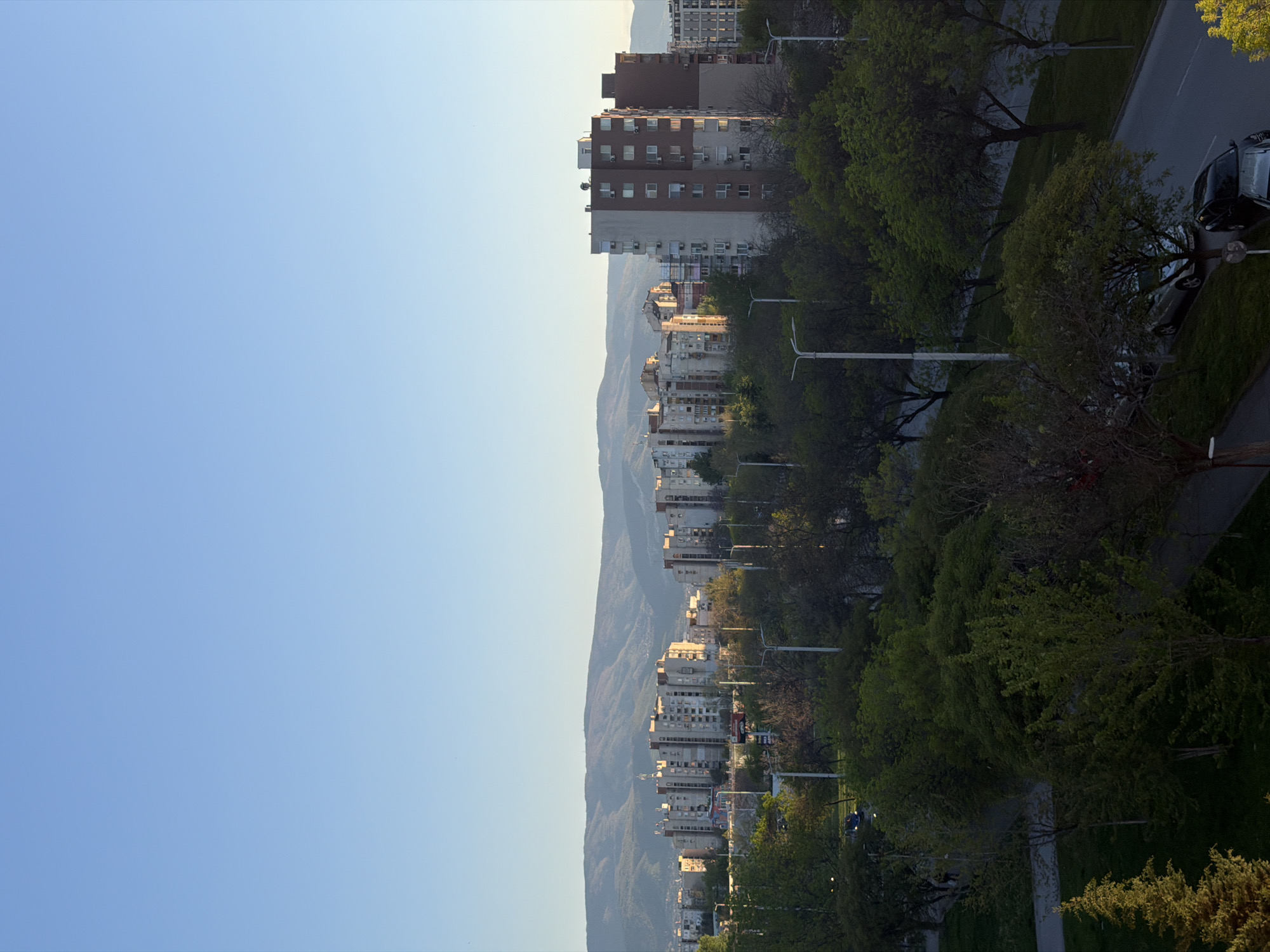 This image captures a serene urban landscape in Plovdiv, Bulgaria, bathed in the soft, warm light of either early morning or late afternoon. The scene is dominated by a clear, light blue sky that fills the upper portion of the frame, indicating pleasant weather.
Stretching across the mid-ground is a long line of multi-story buildings, primarily residential apartment blocks, showcasing varied architectural styles with numerous windows, balconies, and visible air conditioning units. Behind these buildings, a majestic mountain range rises in the distance, its peaks appearing somewhat hazy, adding depth to the scene.
In the foreground, on the right side, a lush, tree-lined area is visible, likely a street or park. Numerous deciduous trees display fresh, vibrant green foliage, suggesting spring or early summer. Several modern streetlights are positioned along this area. A paved road or pathway runs along the far right, where a dark-colored car is partially visible, parked.
The lighting creates a peaceful atmosphere, with the gentle sunlight illuminating the building facades and the distant mountains with a soft glow, while shadows are long but not deep. No people or specific activities are discernible in the tranquil setting. There is no visible text in the image.