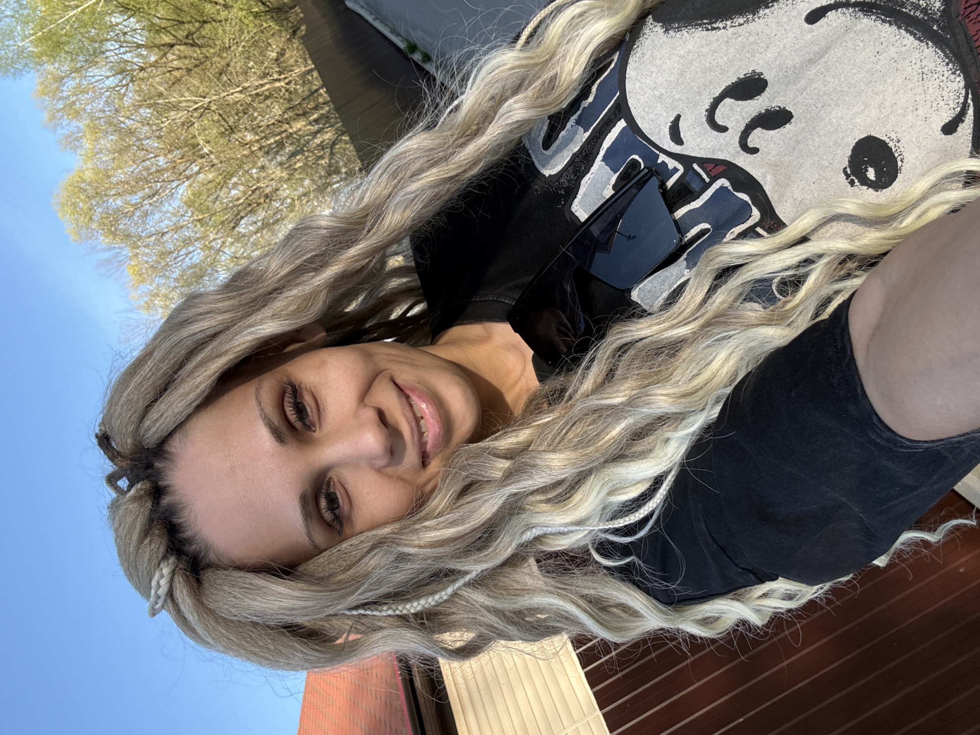 A fair-skinned woman with blond, braided hair is facing the camera with a slight smile. She is wearing a black T-shirt with a white cartoon graphic and sunglasses. The background consists of a blue sky, tree branches with green leaves, a red building, and a brown wooden surface. The location is likely outdoors during daylight. The city of Rusanovo, Russia, cannot be confirmed from the image.