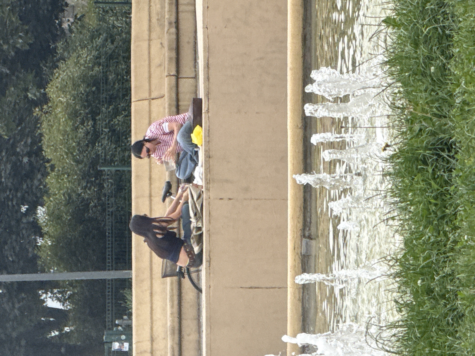 The image displays an outdoor scene featuring two people alongside a multi-jet fountain, likely in a park or public garden, under bright daylight. The photograph is oriented vertically, but depicting a horizontal scene.
On the left side of the frame, a person with dark hair, wearing sunglasses, a red and white striped shirt, and blue jeans, is seen in a crouching or bent-over position. A yellow item, possibly a piece of clothing or a bag, is near their feet, and they appear to be interacting with a small dark object near their hand.
To their right, a second person with long, dark hair is seated on a bicycle, which rests against a light-colored concrete or stone barrier. This person is wearing a dark top and light-colored shoes, and appears to be holding a small white object in their hand.
The barrier separates the individuals from a long fountain, where numerous jets of water erupt vertically, creating a dynamic watery surface. Beyond the fountain, on the far right, a strip of vibrant green grass is visible.
In the background on the left, behind the first person, there is a dense expanse of green trees and foliage, suggesting a natural or heavily landscaped environment. The bright lighting indicates a sunny day. No legible text is visible in the image. The scene conveys a moment of relaxation or activity in a public green space.