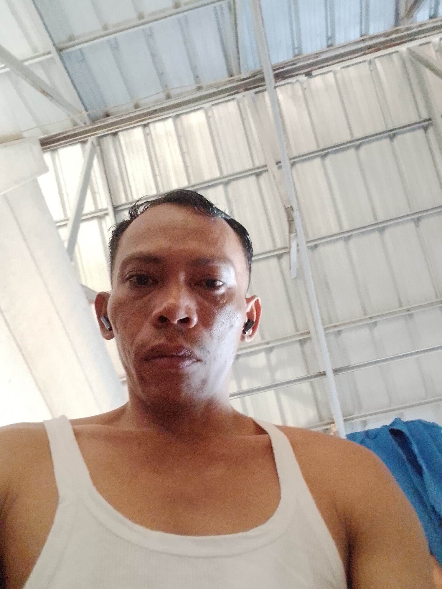 A low-angle, medium shot captures a man from the chest up, looking directly at the camera. He is wearing a white tank top and has dark hair, styled neatly. The man has earbuds in both ears. His face is illuminated, suggesting the sun is overhead. The background consists of a corrugated metal roof supported by a metal frame. The scene appears to be outdoors, possibly a workshop or covered area in Bukit Ketok, Indonesia.