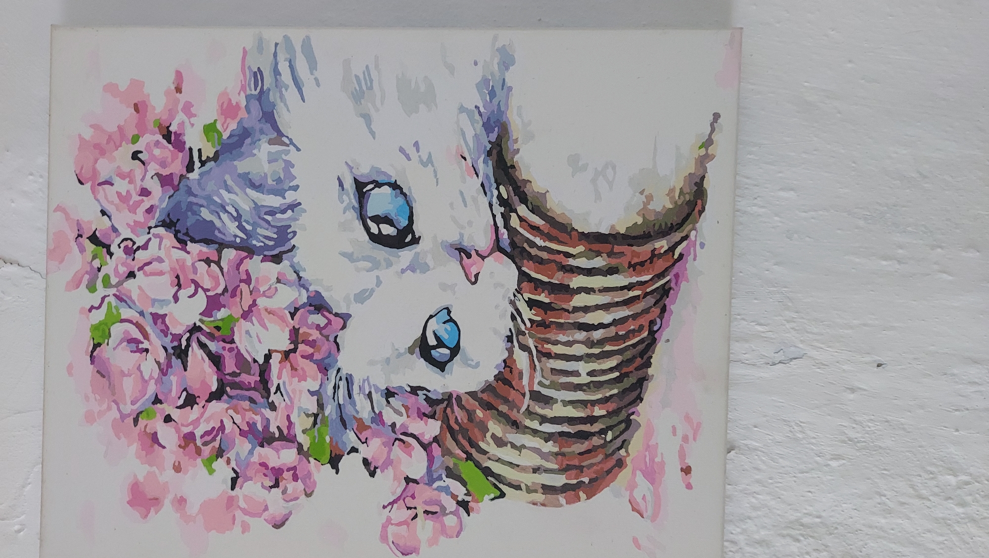 A framed painting of a white cat wearing a floral headdress is displayed on a textured white wall. The cat has large blue eyes and a pink nose. It is resting its head on a brown and white striped cylindrical object. The floral headdress is composed of pink and purple flowers. The painting is rendered in a stylized, illustrative manner.