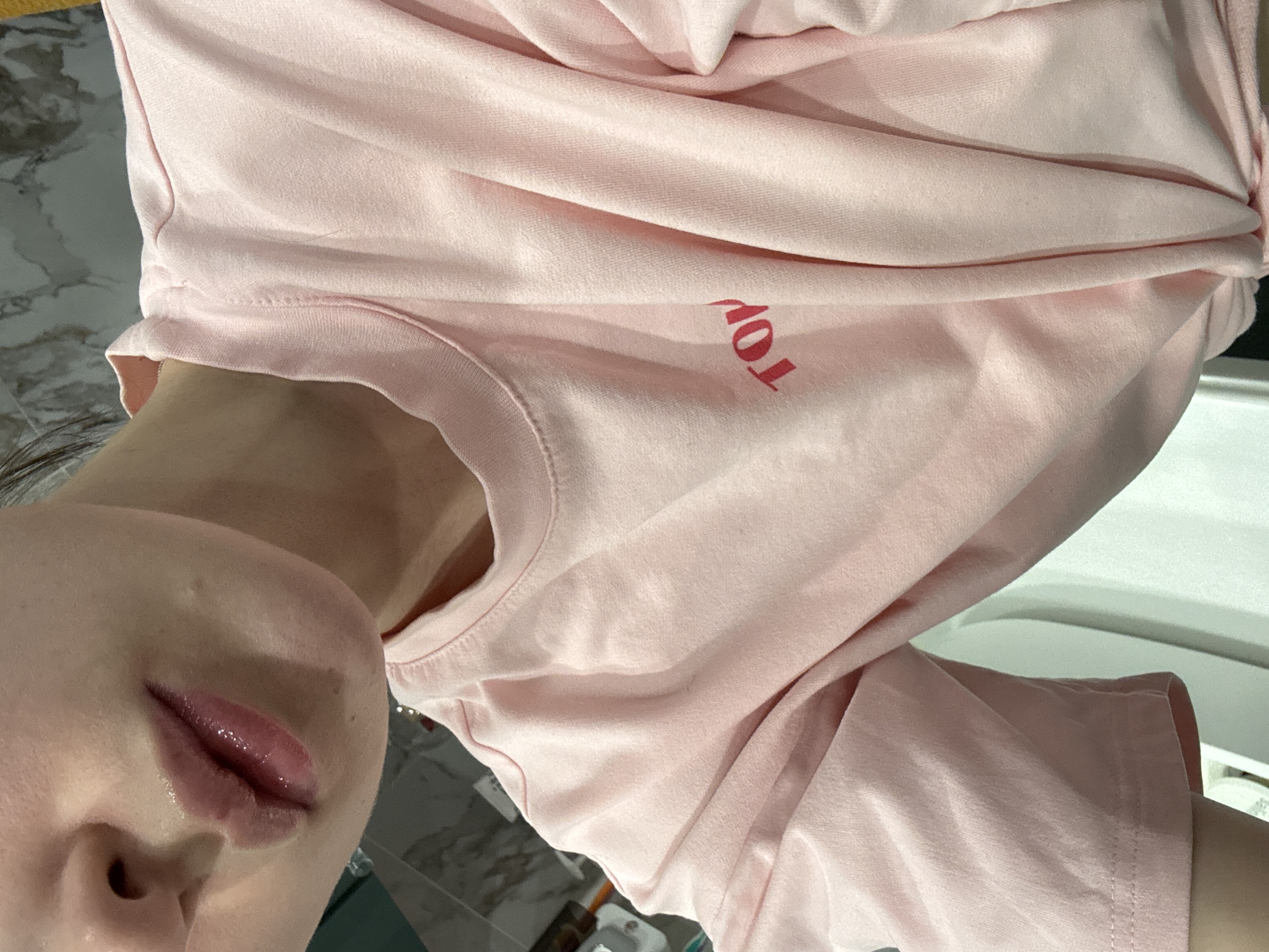 The image shows a close-up view of a person wearing a pink t-shirt. The word "TOP" is visible on the shirt. Part of the person's face, including the nose and lips, is in the frame. The background is blurry and shows what appears to be a bathroom counter with toiletries and tiles. There is no information to verify its precise location in Tomilino, Russia.