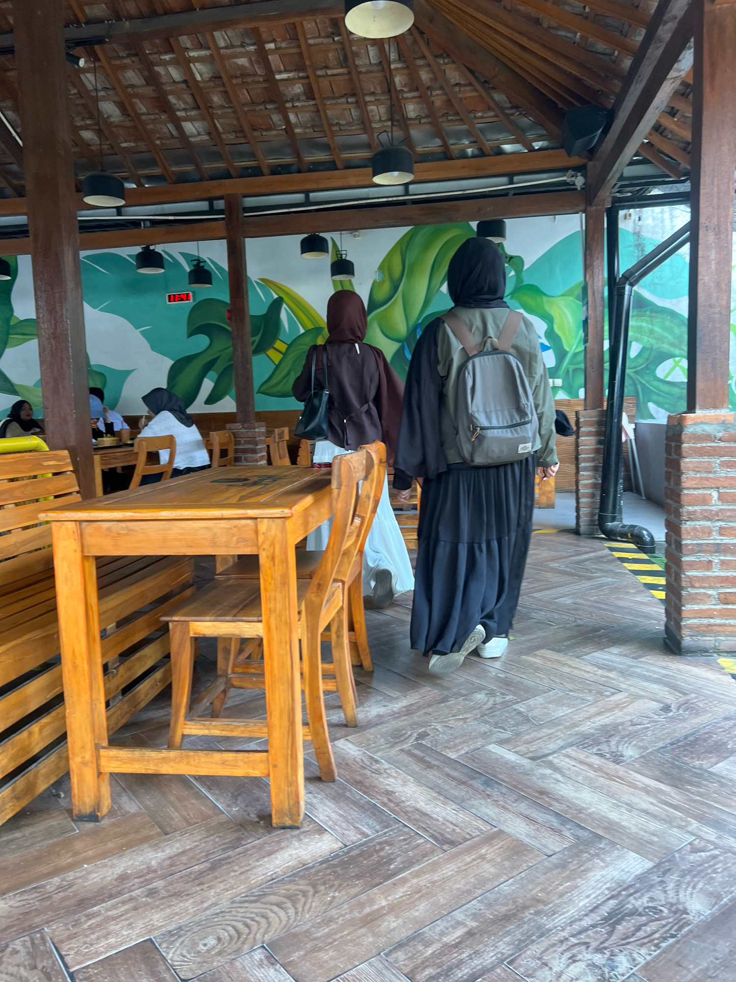 The image captures an indoor dining area with an open-air, rustic aesthetic, likely located in Yogyakarta, Indonesia. The scene features wooden tables and benches, a tiled floor with a wood-like pattern, and a ceiling constructed of exposed wooden beams and terracotta tiles. A mural depicting large green leaves adorns the back wall.
Two individuals wearing hijabs are the primary subjects. The person in the foreground, walking away from the camera, is wearing a black hijab, a khaki-colored jacket, and a long black skirt. They are also carrying a gray backpack. To their left, another person in a dark brown hijab and light-colored clothing is visible. In the background, several other patrons are seated at tables, some wearing hijabs.
The overall atmosphere suggests a casual dining establishment, possibly a cafe or restaurant, with natural light filtering in and a blend of traditional and modern design elements. The presence of a digital clock showing "11:41" indicates the time of day.