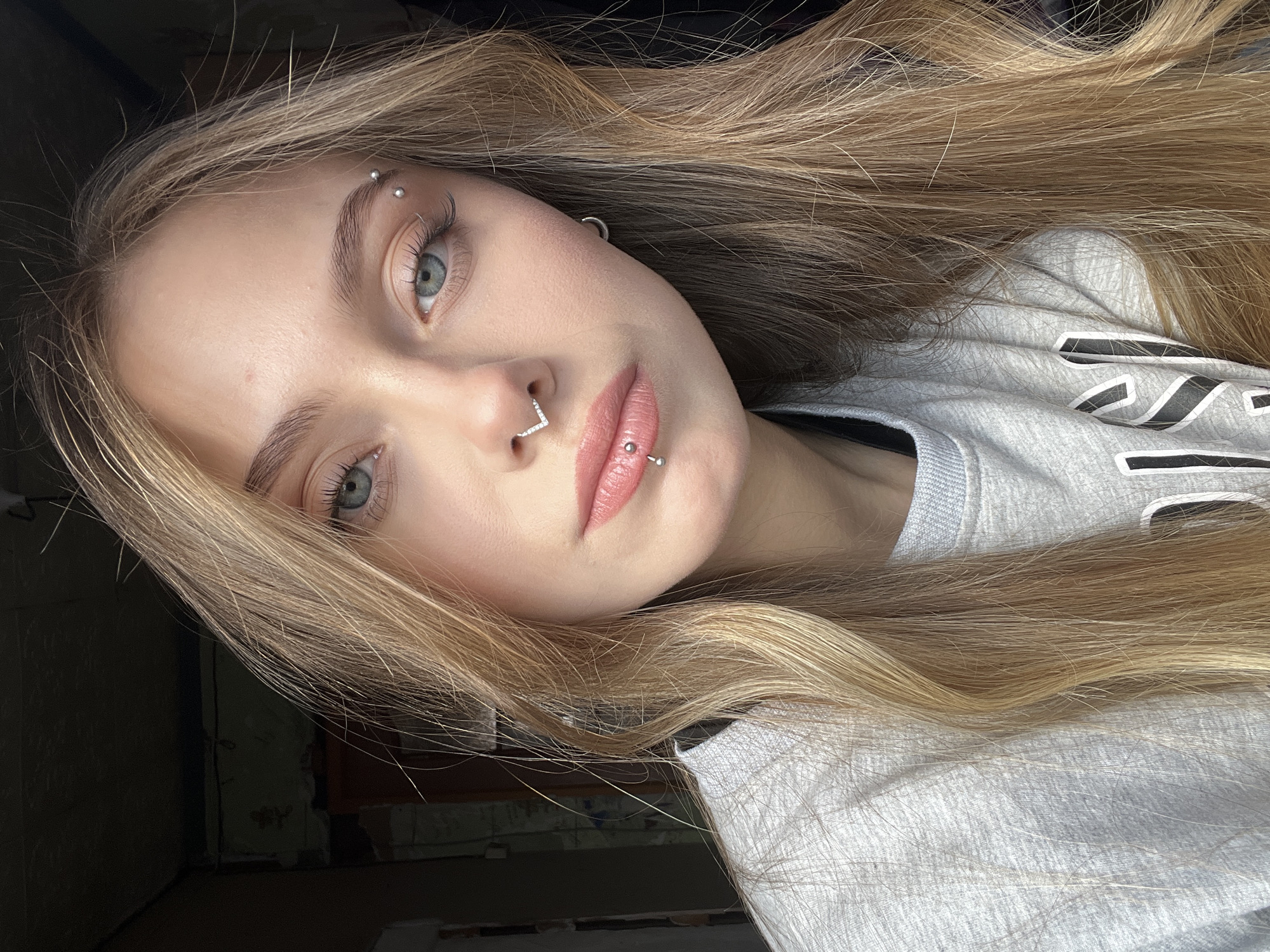 A close-up portrait features a young person with long, wavy blonde hair and visible darker roots, fair skin, and light blue-grey eyes. The individual has distinct facial piercings, including an eyebrow piercing with two silver beads, a silver V-shaped septum ring adorned with small jewels, and a philtrum (Medusa) piercing with a small silver stud. Their lips are lightly covered with a soft pink lip gloss or lipstick, and their eyebrows appear well-defined.
The person is wearing a light grey crewneck sweatshirt. Visible white and dark grey lettering on the chest includes the letters "NI," characteristic of the "NIKE" brand logo.
The image is a portrait, likely a selfie, captured with natural light illuminating the subject's face, suggesting it was taken during the daytime. The background is dimly lit and largely out of focus, appearing to be an indoor setting. On the left side, textured wall surfaces, possibly unfinished or aged, are visible in shadow. The subject maintains a neutral, composed expression while looking directly at the camera.