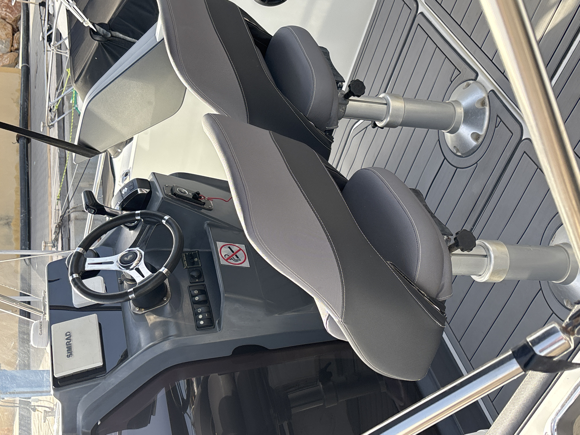 The image shows the helm of a modern boat docked at a marina in Urbanización Cabo Roig, Spain. The helm features a SIMRAD navigation screen and a steering wheel. Two adjustable boat seats, upholstered in grey and dark grey fabric with white stitching, are positioned in front of the helm. The seats are mounted on polished metal pedestals. The boat's deck is covered with a grey non-slip mat. In the background, other boats and a pier can be seen, with a rocky shoreline and buildings of Urbanización Cabo Roig in the distance. The scene is set in daylight, with sunlight casting shadows on the deck.