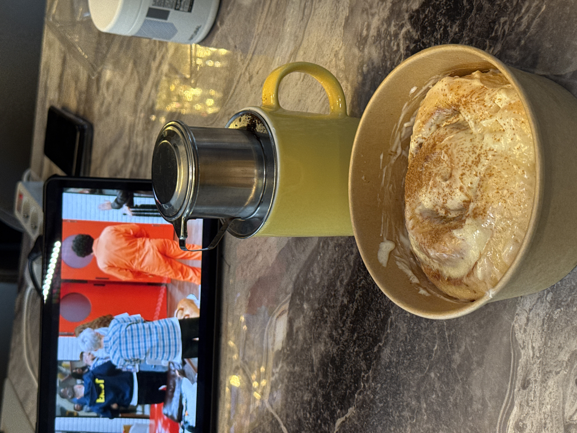 The image shows a tabletop scene. A yellow mug with a metal coffee filter sits next to a bowl of what appears to be cinnamon rolls with icing. To the left, a tablet is displaying a video scene of a person in an orange outfit and another person in a blue plaid shirt. Other objects include a white container and a small black object near the tablet. The setting appears to be a kitchen or dining area, with a marble-patterned countertop. The location in the city of Unknown, Unknown is not determinable from this image.