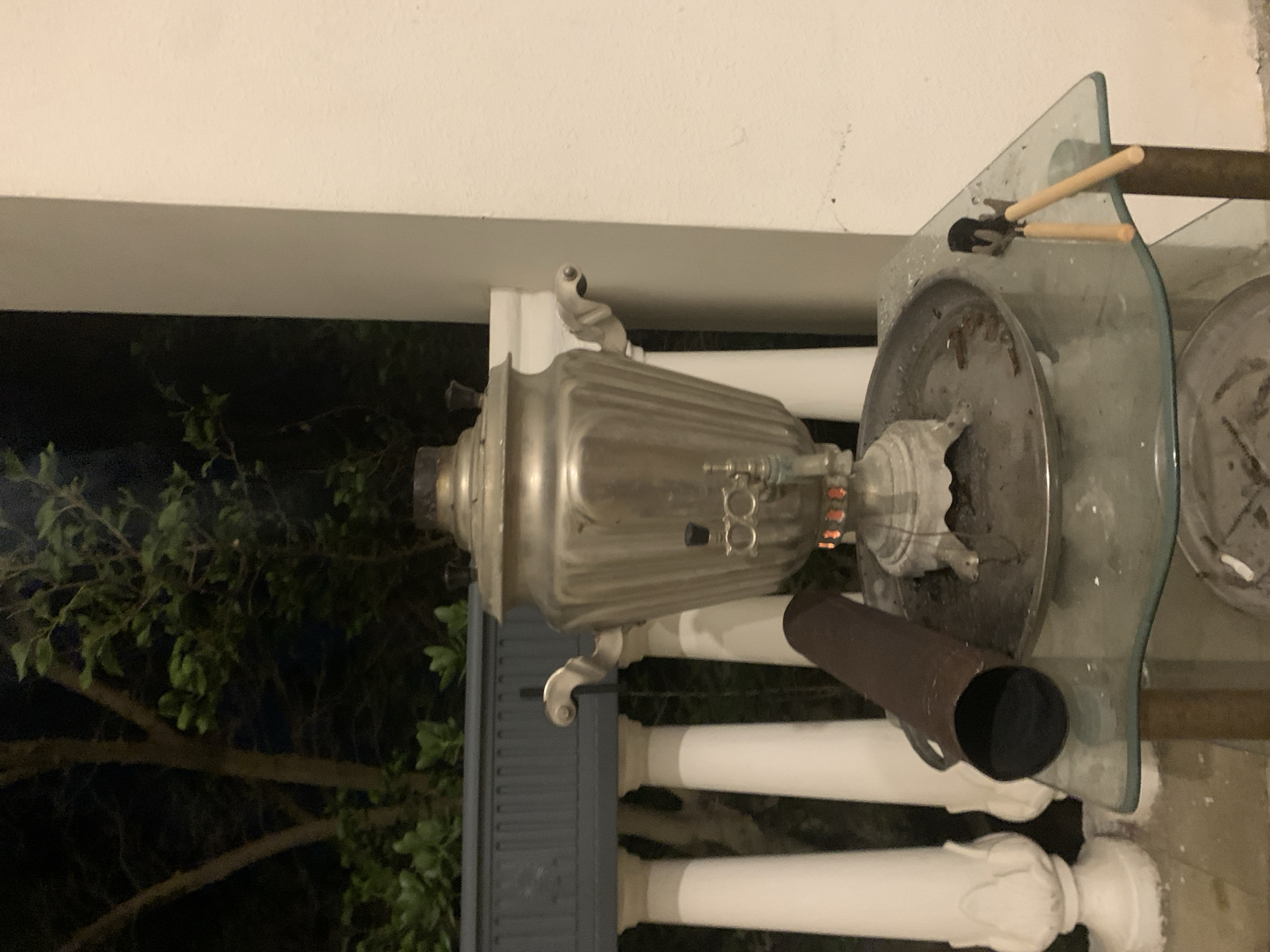 The image depicts an outdoor setting at night, with foliage visible in the background. The main subject is a large, ornate, metal samovar, a traditional Russian vessel for heating and boiling water. The samovar is silver-colored and has a ribbed body with decorative handles and spouts. It appears to be placed on a glass surface, which is supported by a metal structure.
To the right of the samovar, there is a dark, cylindrical object, possibly a chimney or exhaust pipe, extending from the samovar. Also on the glass surface are two small gardening tools with wooden handles, one black and the other brown. The background shows a dark, leafy tree, suggesting a garden or outdoor area. The overall scene appears to be a static display rather than an active event. The lighting suggests it is nighttime, and there are no people visible in the image. The setting appears to be a patio or balcony area, with white, decorative railing posts visible in the foreground. The environment is likely in Khazar Raion, Azerbaijan, as indicated by the location context. There is no visible text in the image.