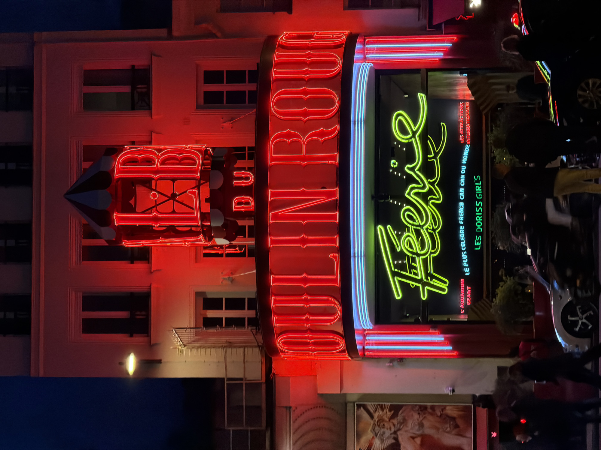 The image depicts the Moulin Rouge in Paris, France, at night. The building's facade is brightly illuminated with red neon lights spelling "Moulin Rouge" vertically on the right, with a smaller, illuminated windmill feature above on the left. The word "Féerie" is displayed in green neon. There's a sign saying "Le plus celebre french can can du monde". People are walking near the building. A car is parked on the right, partially obscuring the view. The windows of the building are visible above the signage.