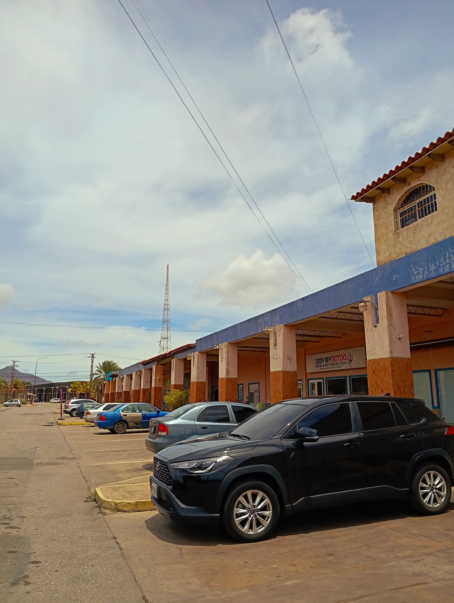The image depicts an outdoor commercial street scene, identified as being in the city of Juan Griego, Venezuela, under a partly cloudy daytime sky.
In the foreground, a paved parking lot occupies the lower portion of the frame, showing faded yellow parking space delineations. A black Toyota Corolla Cross SUV is prominently parked on the right. Behind it, a light blue sedan, a damaged blue sedan with its rear passenger door panel visibly absent, and a silver sedan are parked in sequence along a yellow curb. Additional vehicles are partially visible further down the street on the left.
The midground features a long, single-story building complex. Its architecture includes light-colored walls, a distinct blue horizontal band along the upper facade, and a red-tiled roof. The structure is supported by multiple pillars, creating an open, covered walkway. One clearly visible business sign reads "TOMY REVTATTOO. TATTOO & BODY PIERCING STUDIO." The building's end section on the right has an arched window fitted with security grates on an upper level. Palm trees and utility poles with overhead power lines are present to the left of the building complex.
In the background, a tall, lattice-structured telecommunications or radio tower stands prominently behind the buildings. Distant, hazy mountains or hills are visible on the horizon to the far left. The sky is a mix of light blue and scattered white clouds. No individuals are discernible within the scene.